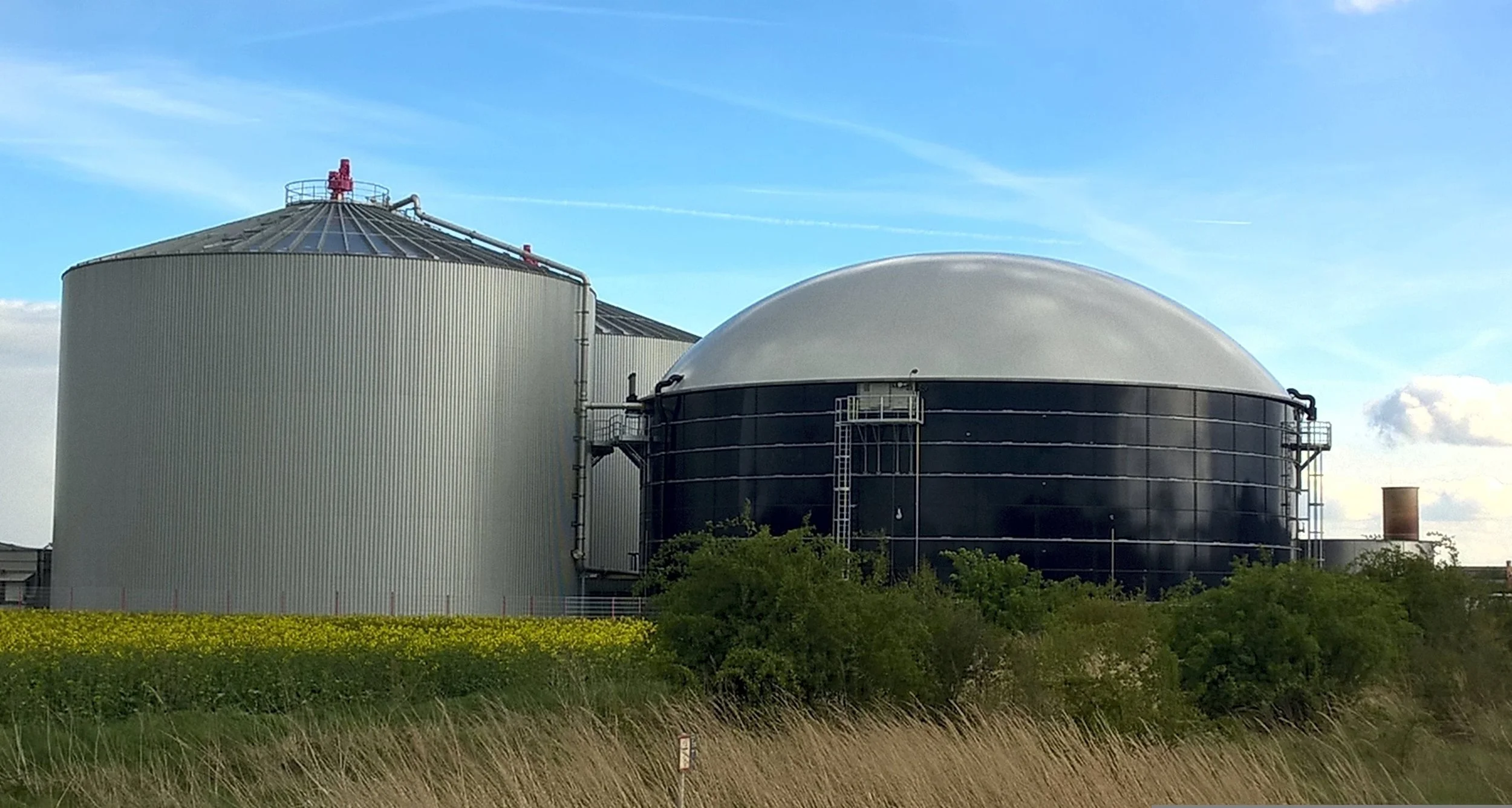 Large Scale Anaerobic Digestion Plant including one large gray cylindrical tank and one smaller black spherical tank, are situated outdoors with green bushes and a yellow flower field in the foreground and a partly cloudy blue sky in the background.