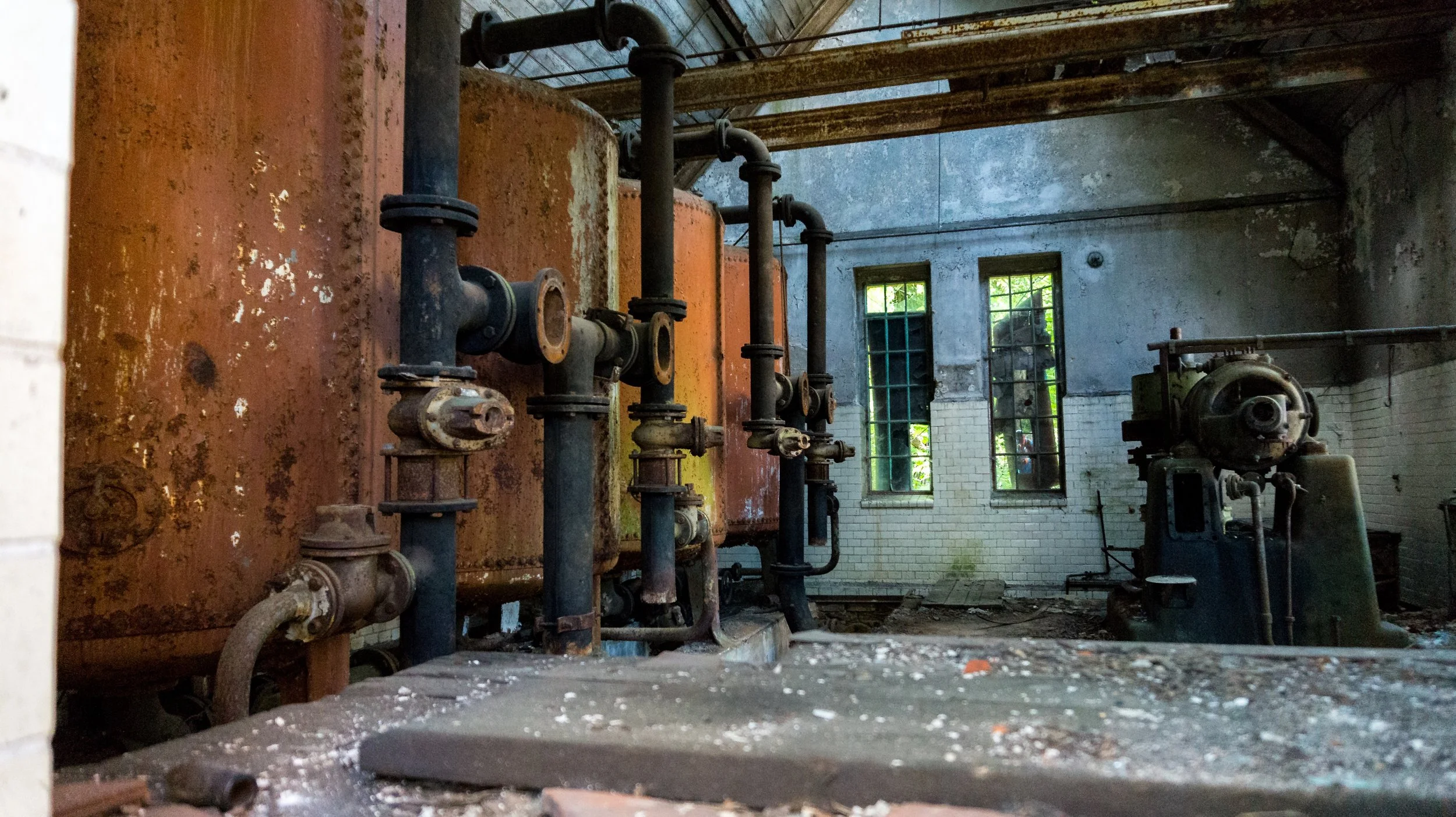 Old, rusted industrial boiler system with pipes and machinery inside an abandoned building with tall windows and peeling walls.