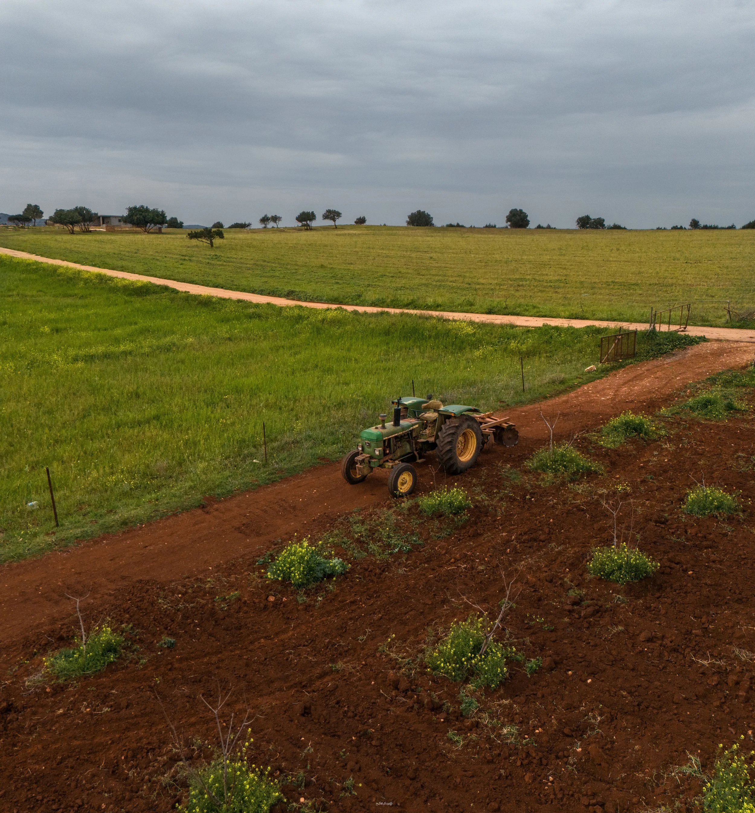 A landscape scene with a dirt path running across green fields under a cloudy sky, some scattered trees, a small building in the distance, and equipment in the foreground.