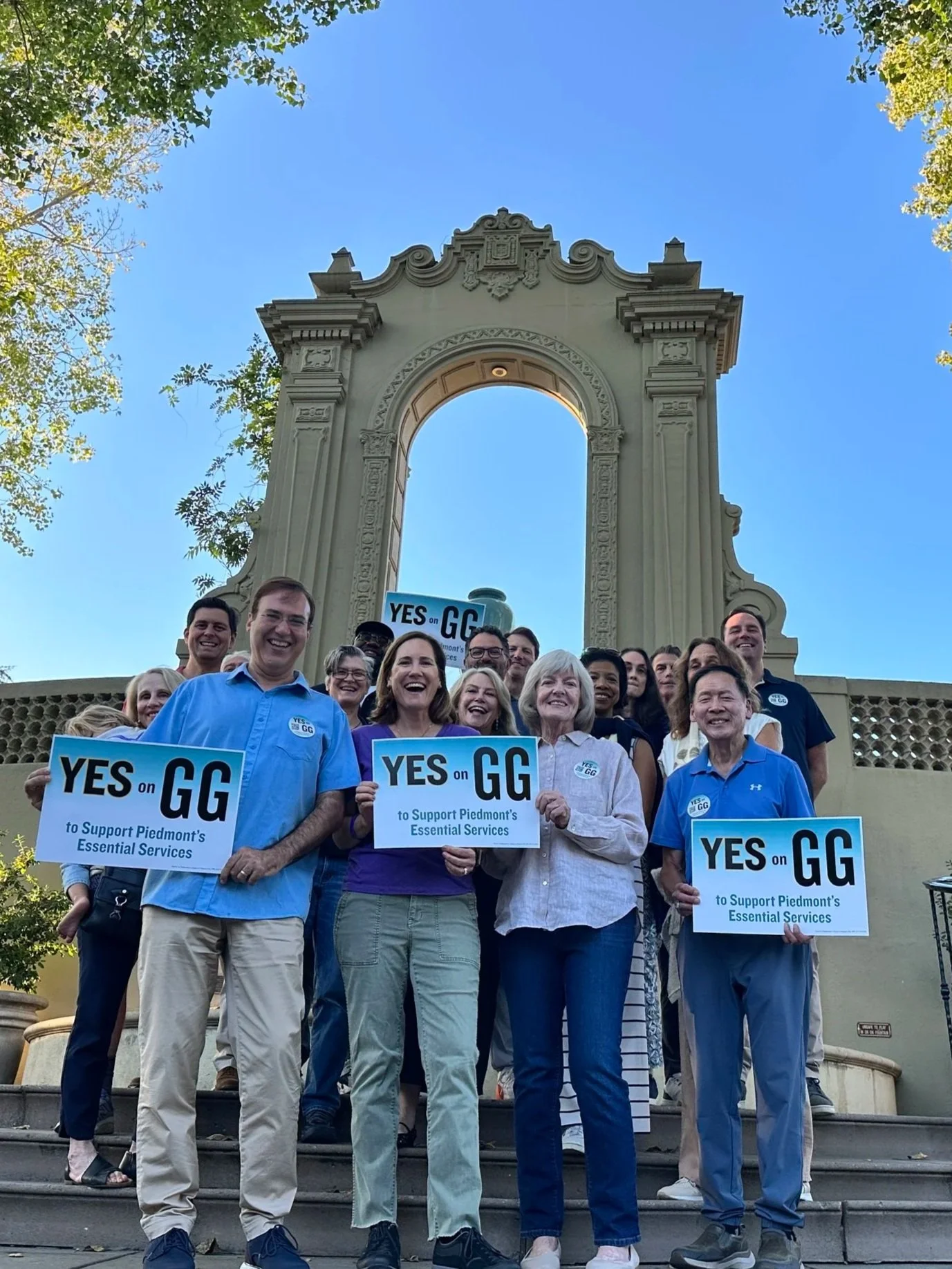 A group of Piedmont Measure GG supporters holding "YES on GG to Support Piedmonts Essential Services" signs in front of the Exedra. Everyone is smiling and may are laughing.