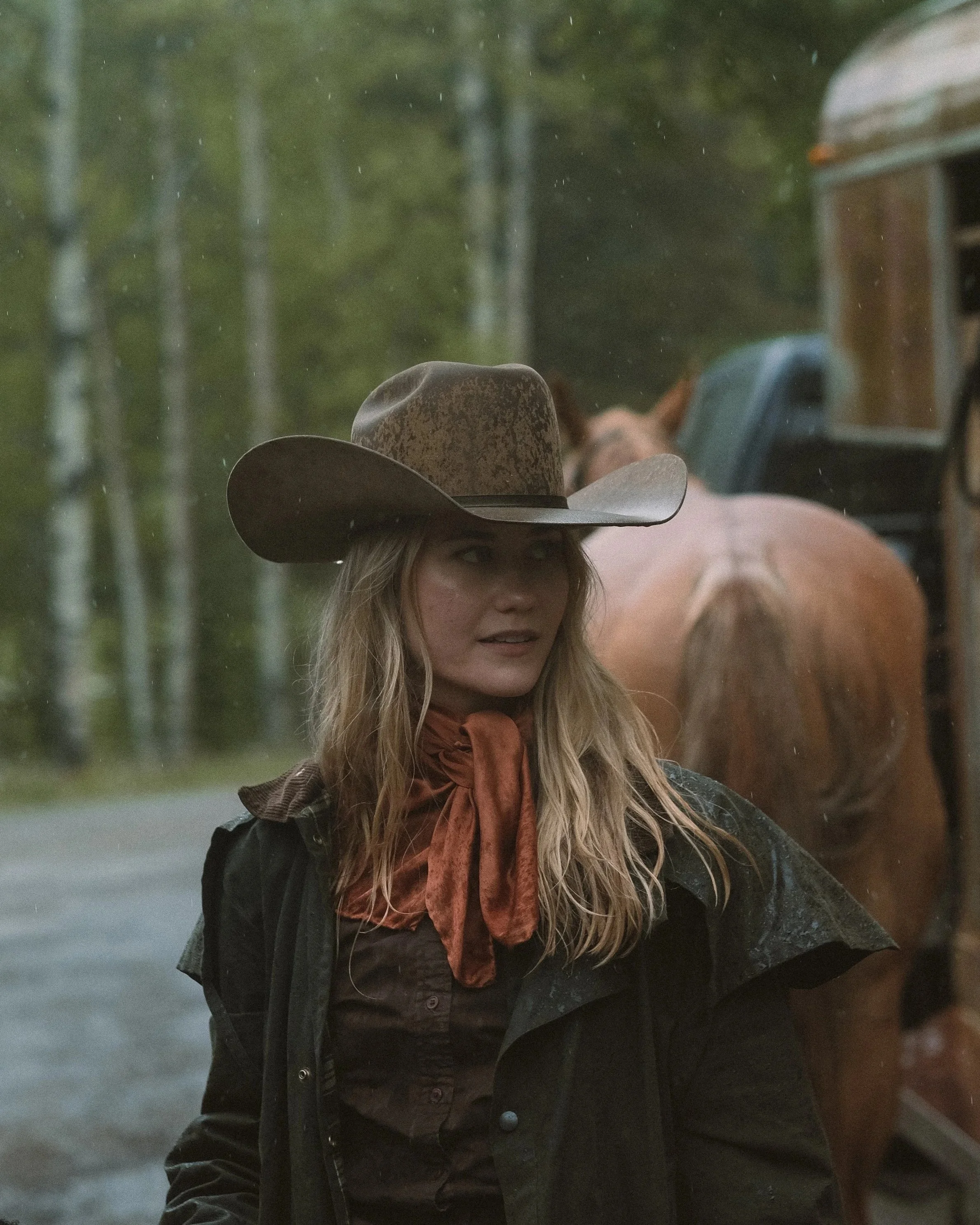 A woman with long blonde hair wearing a cowboy hat and a black jacket, standing outdoors in a wooded area with rain falling.