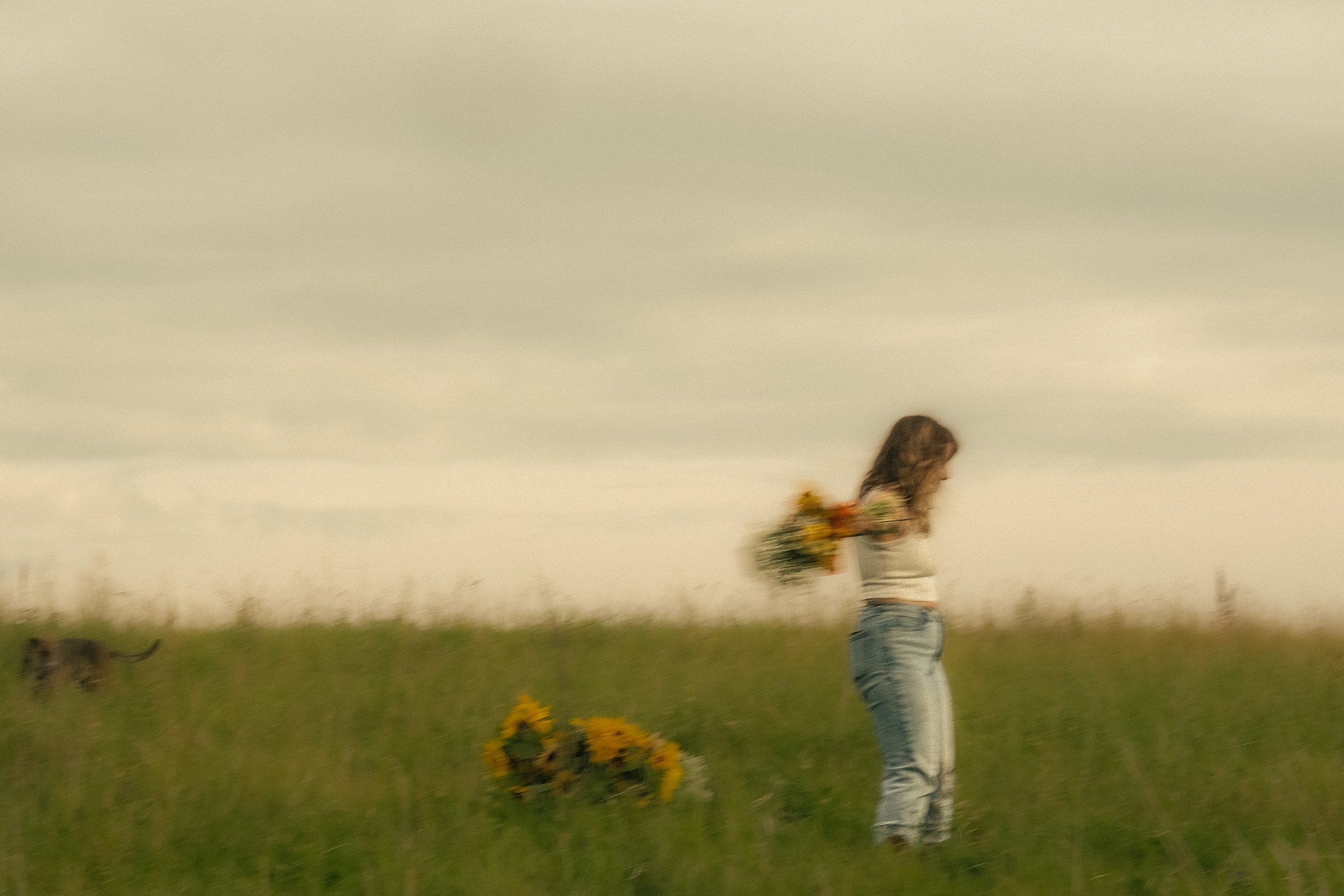 A woman in casual clothing holding a bouquet of flowers and walking in a green field with a large sunflower arrangement nearby.