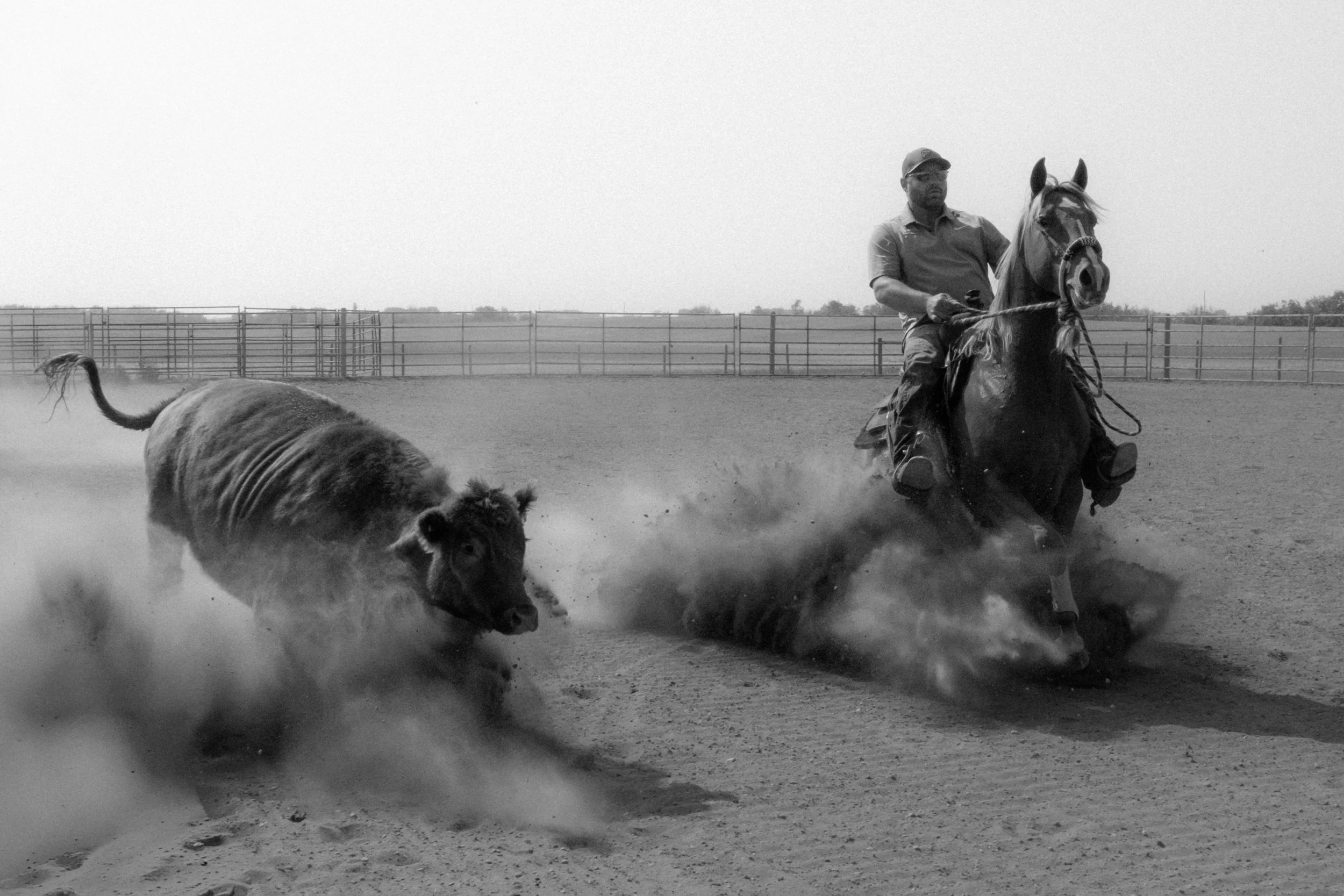 A man riding a horse on a dirt riding arena, with a cattle dog running alongside. The scene is in black and white.
