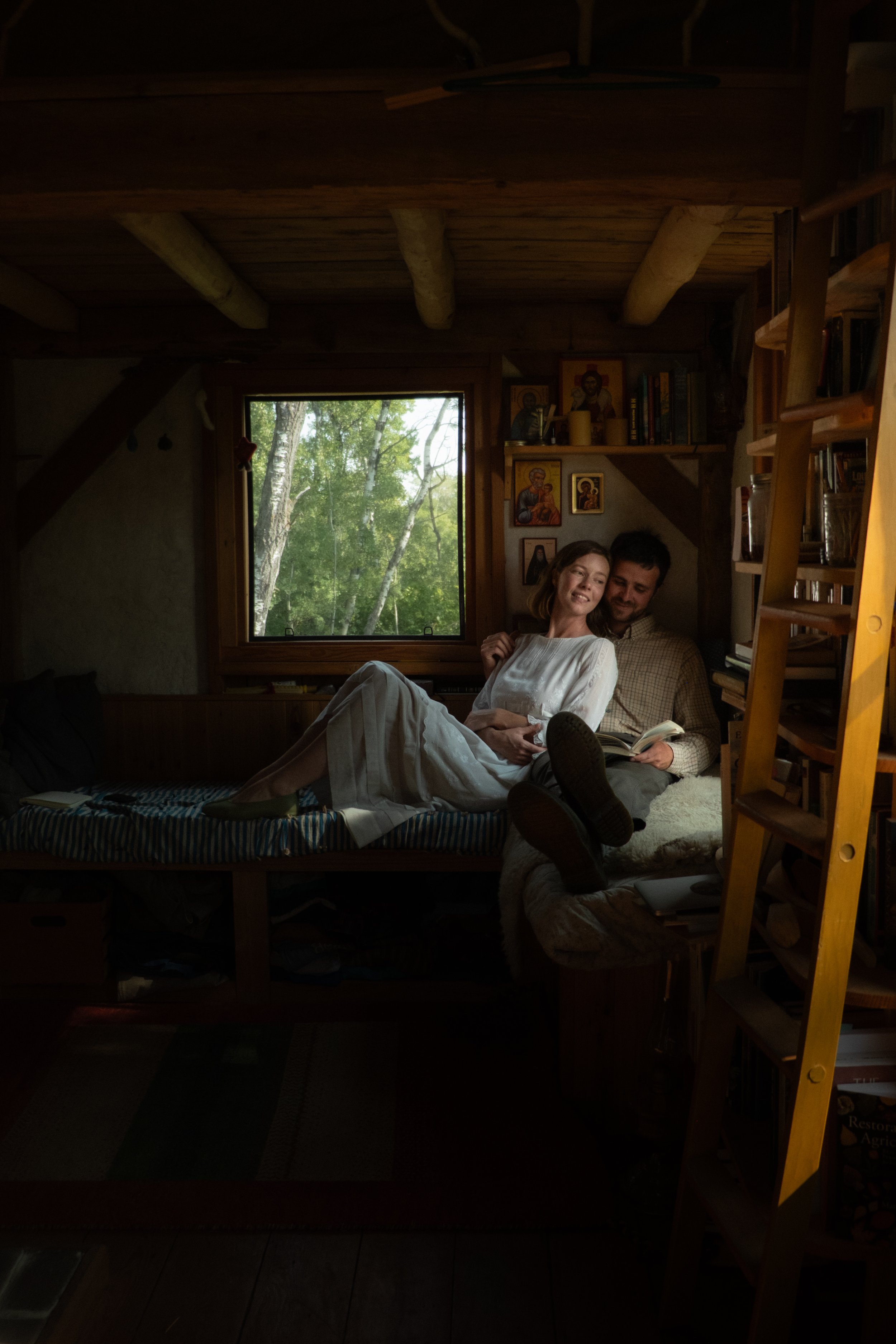 A cozy wooden interior room with a large window showing green trees outside. Two people, a woman and a man, are relaxing on a bed or couch, smiling and enjoying each other's company. The room has bookshelves filled with books and religious icons on the wall.