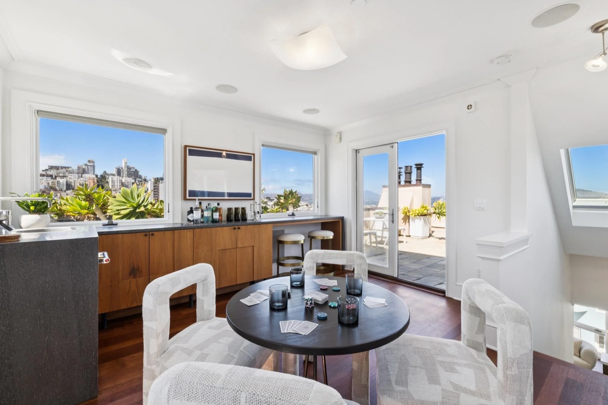 Bright living room with large windows showing city skyline and blue sky, white walls, round black dining table with playing cards and glasses, white woven chairs, and a wooden sideboard with bottles and plants.