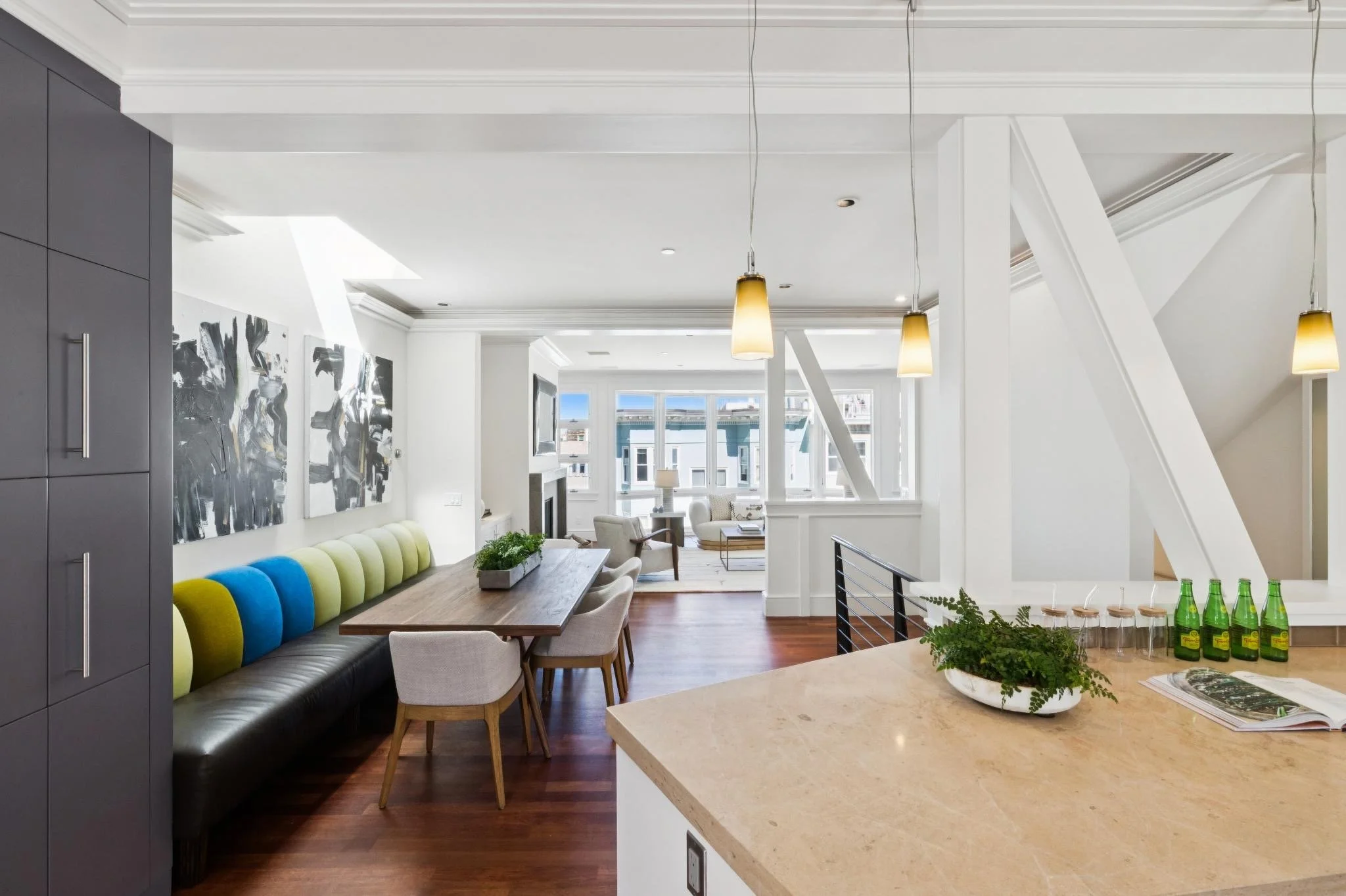 Open-concept living and dining area with large windows, modern artwork, pendant lighting, and a colorful booth seating along a wooden dining table.