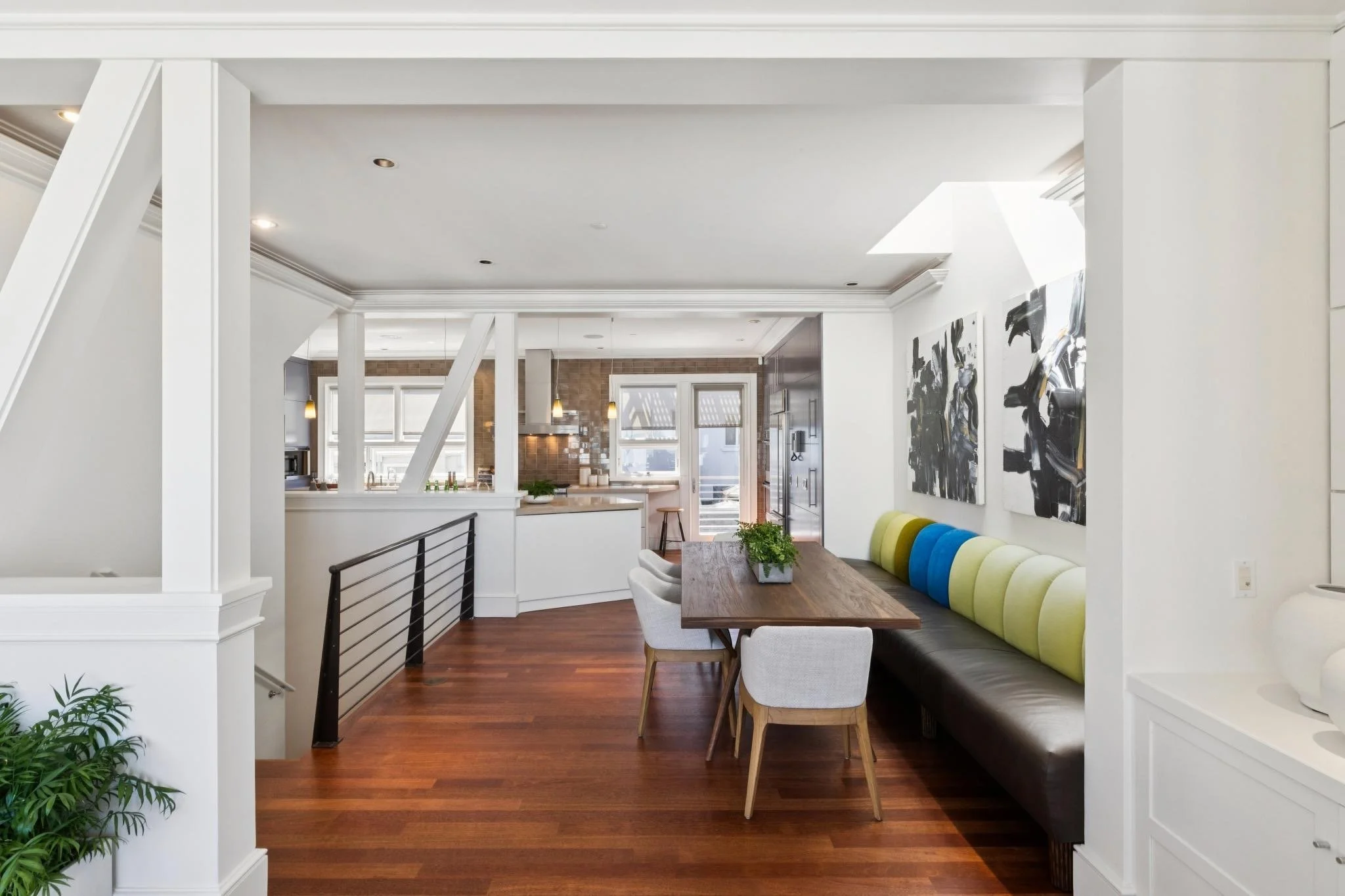 Open-concept living room and kitchen in a modern home with white walls, hardwood floors, abstract wall art, and a built-in seating area with a colorful cushion.