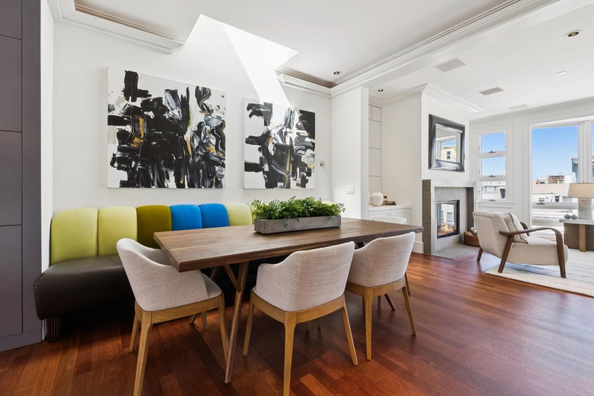 Modern living room with a dining table, beige and colorful chairs, abstract black and white paintings, white fireplace, large windows, and hardwood floors.