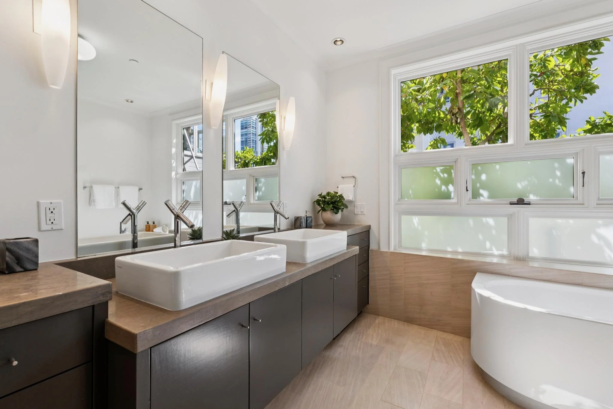 Modern bathroom with double sinks, large mirror, and three large windows showing green trees outside.