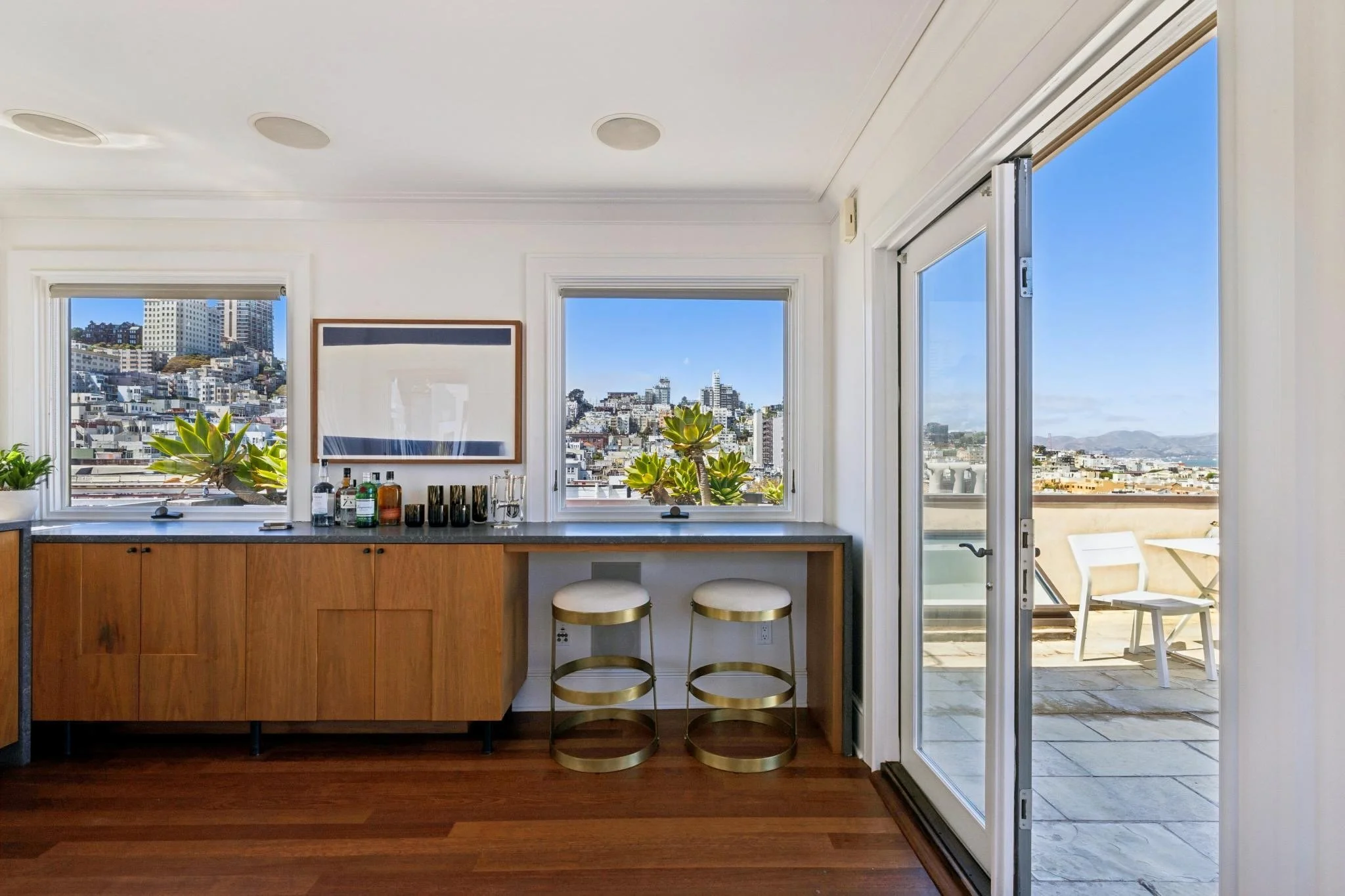 A modern kitchen with a view of a city skyline, featuring large windows, a wooden counter with bottles, plants, and bar stools, and a sliding glass door leading to a balcony with outdoor chairs.