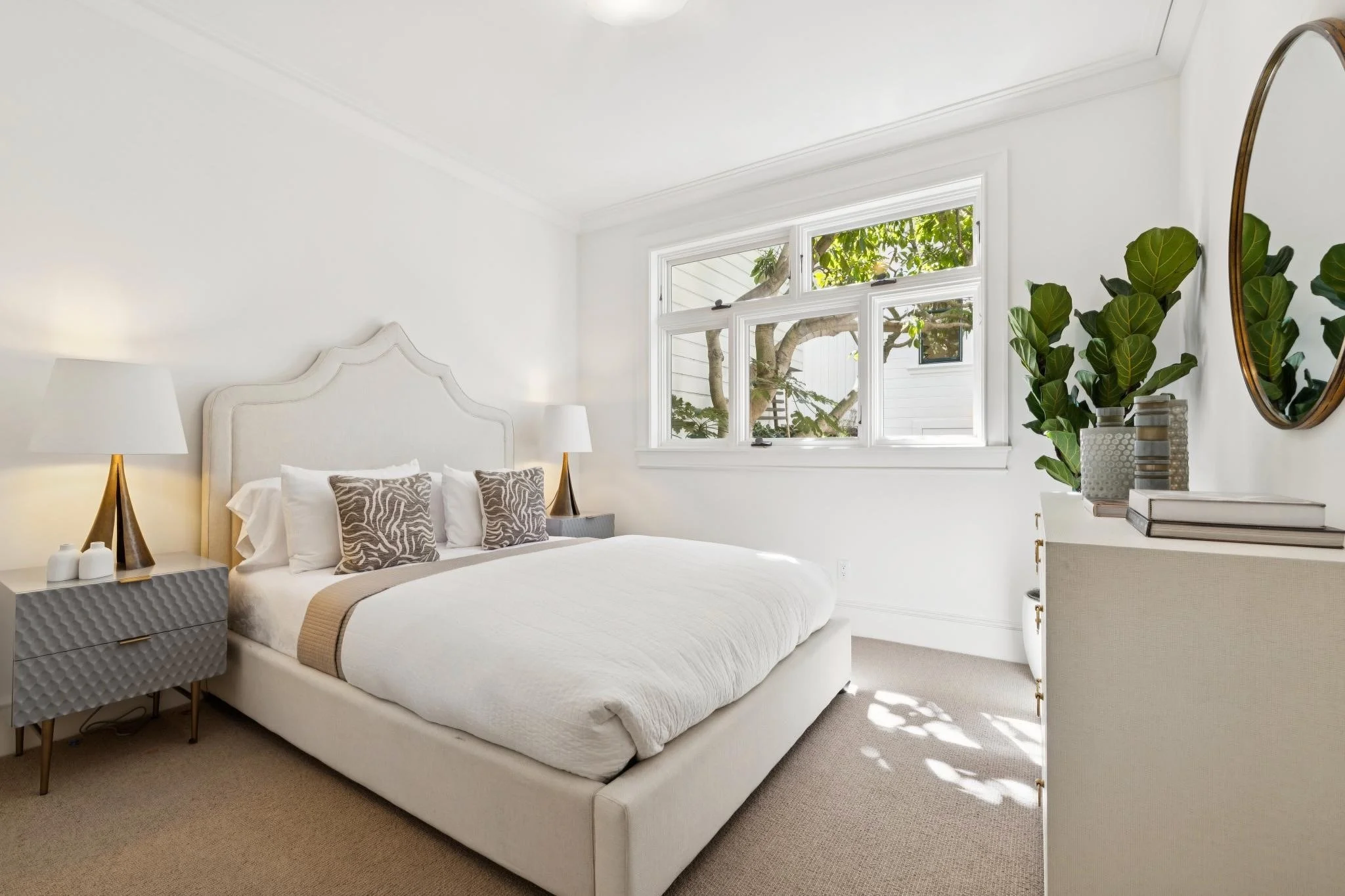 Bright bedroom with white walls, large window showing outdoor greenery, white bed with beige and patterned pillows, bedside table with lamps, a large round mirror, and a dresser with decorative vases.