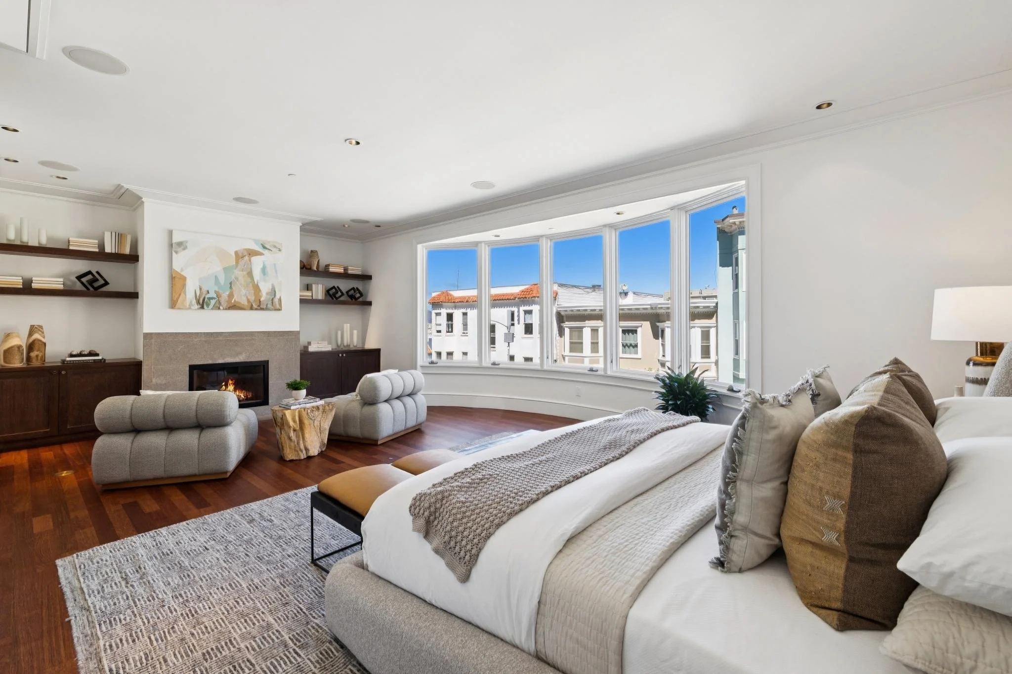 Living room with white walls and large bay window, beige sofa with pillows, modern dark wood bookshelf, abstract wall art, wooden floor, and a white area rug.