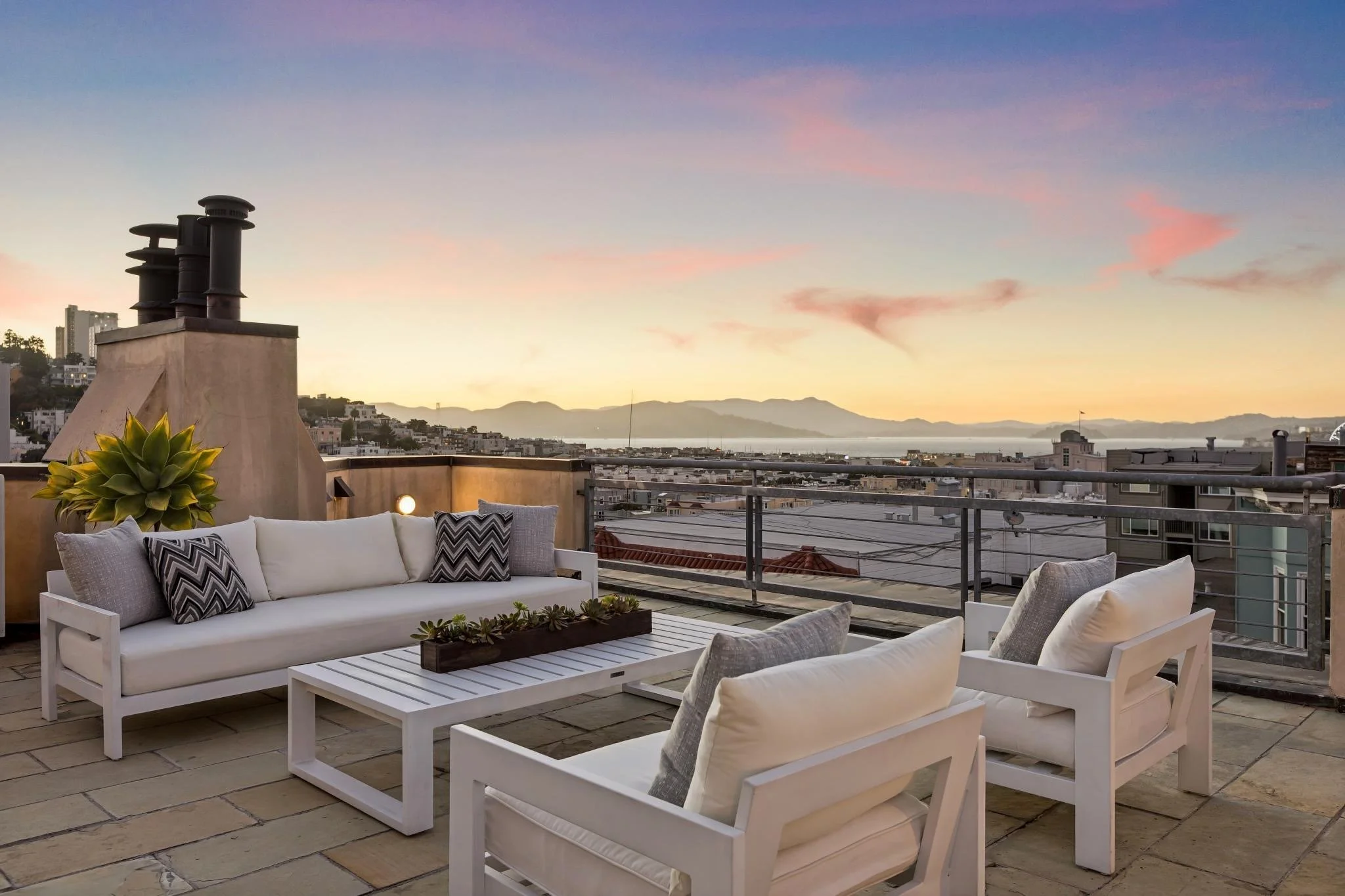 Outdoor patio with white wooden furniture, including a sofa and chairs with cushions, a coffee table with a planter, overlooking a cityscape and distant mountains at sunset.