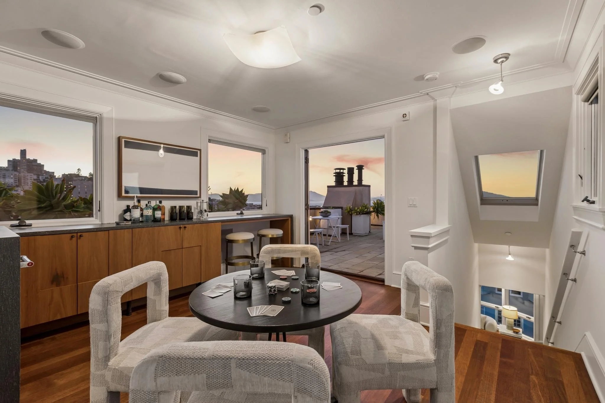 Interior of a room with round dining table, four white upholstered chairs, a wooden sideboard with bottles, and windows showing a sunset sky and cityscape.