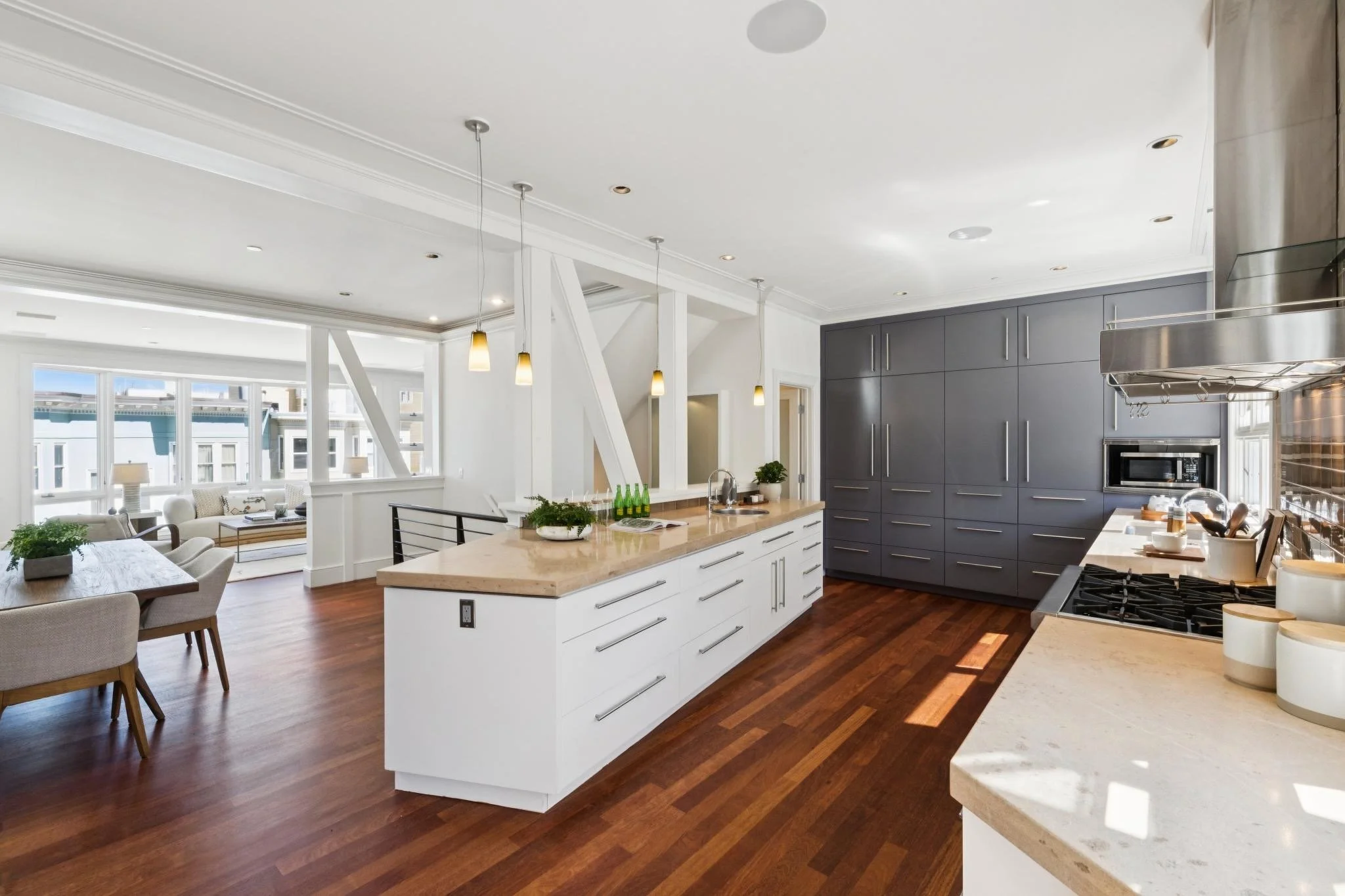 Open-concept kitchen with white cabinetry, dark gray wall cabinets, light granite countertops, and a kitchen island with barstools. The space has hardwood floors, pendant lighting, and large windows providing natural light.