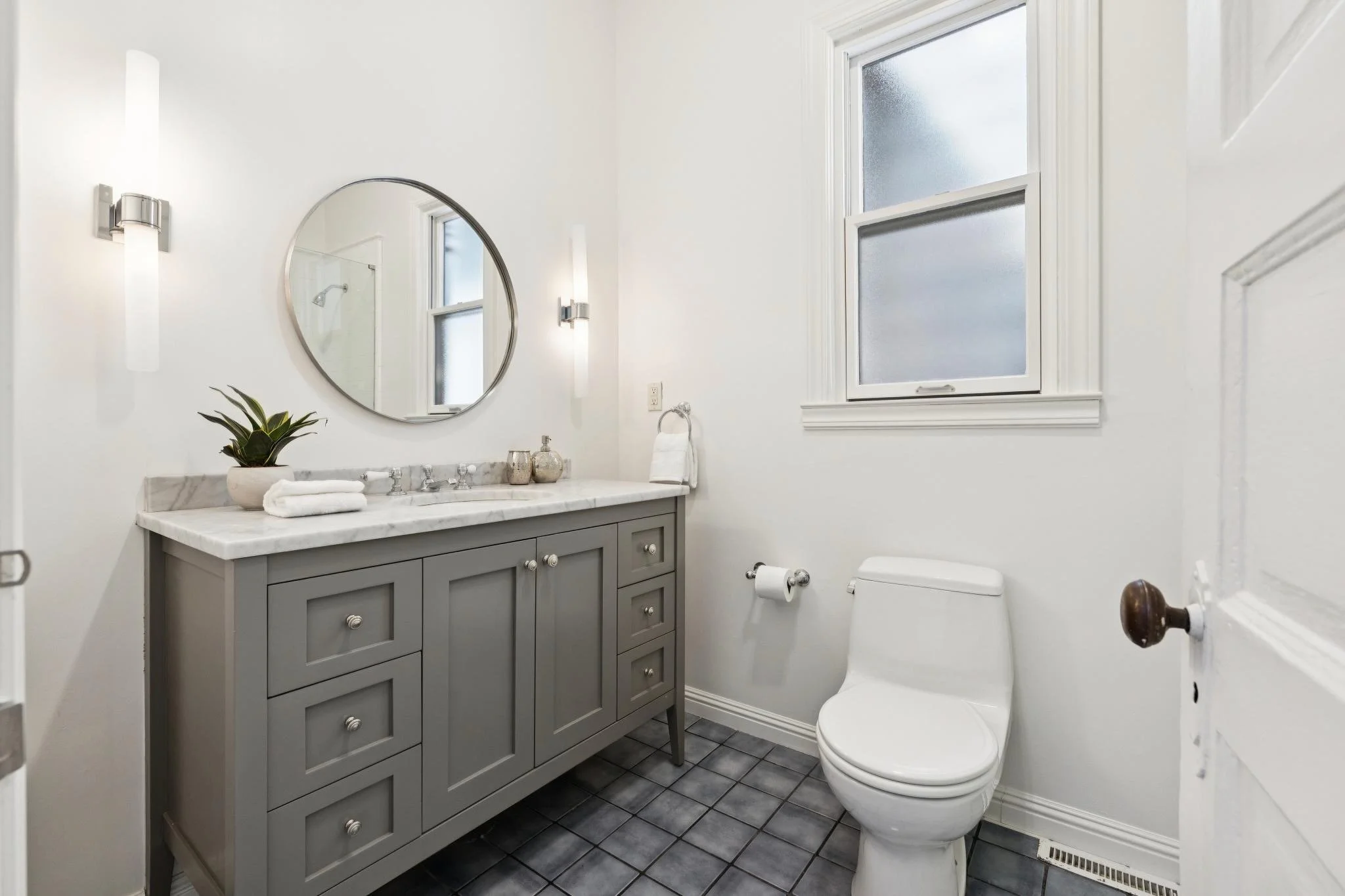 A modern bathroom with a gray vanity, marble countertop, oval mirror, white walls, and a window. There are wall-mounted lights, a plant, towels, and a toilet.