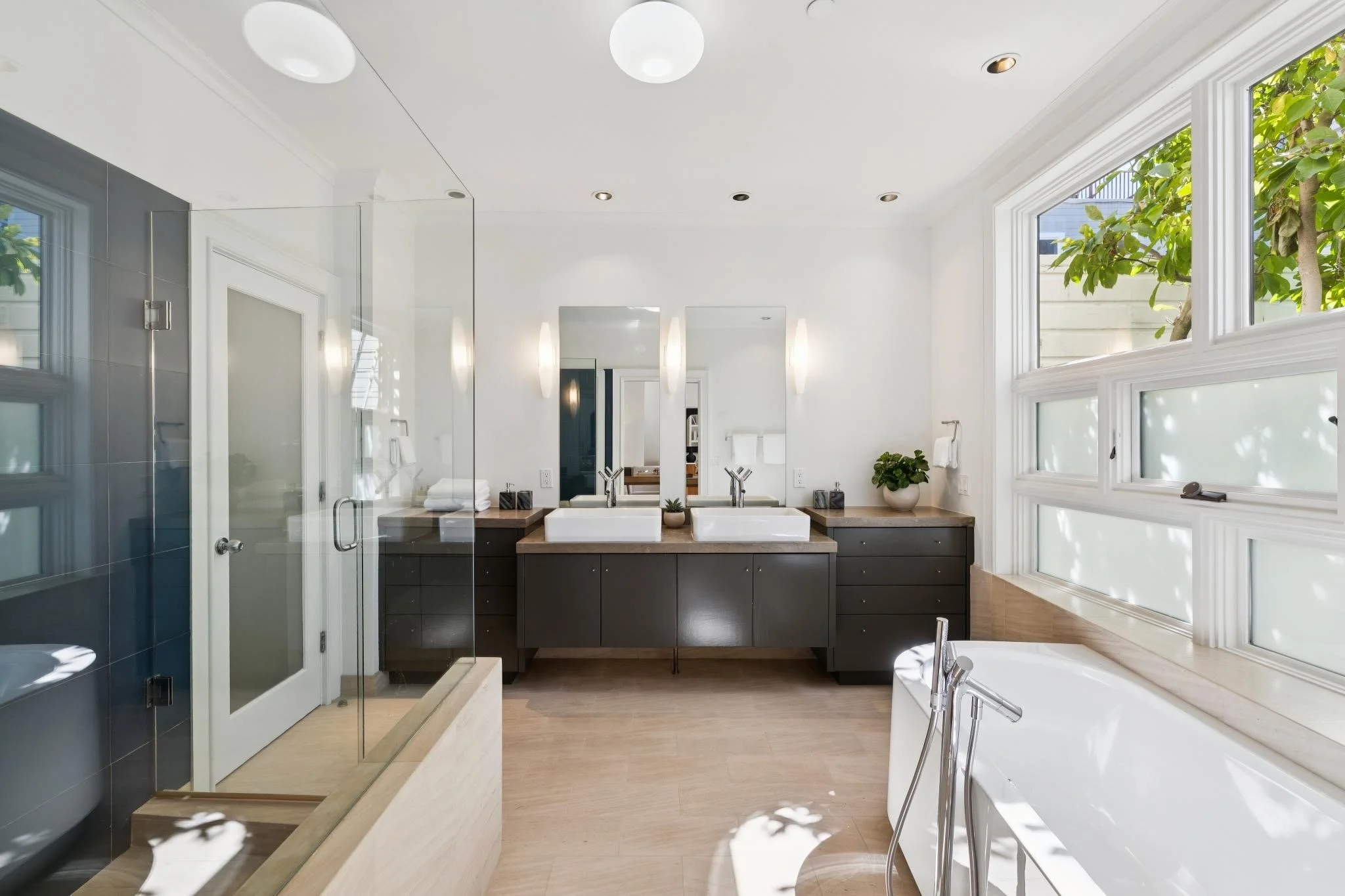 Modern bathroom with large windows, double sinks, and a walk-in shower with glass door. Includes a bathtub, mirror above the sinks, and decorative plants. Natural light streams in, highlighting clean, minimalistic design.