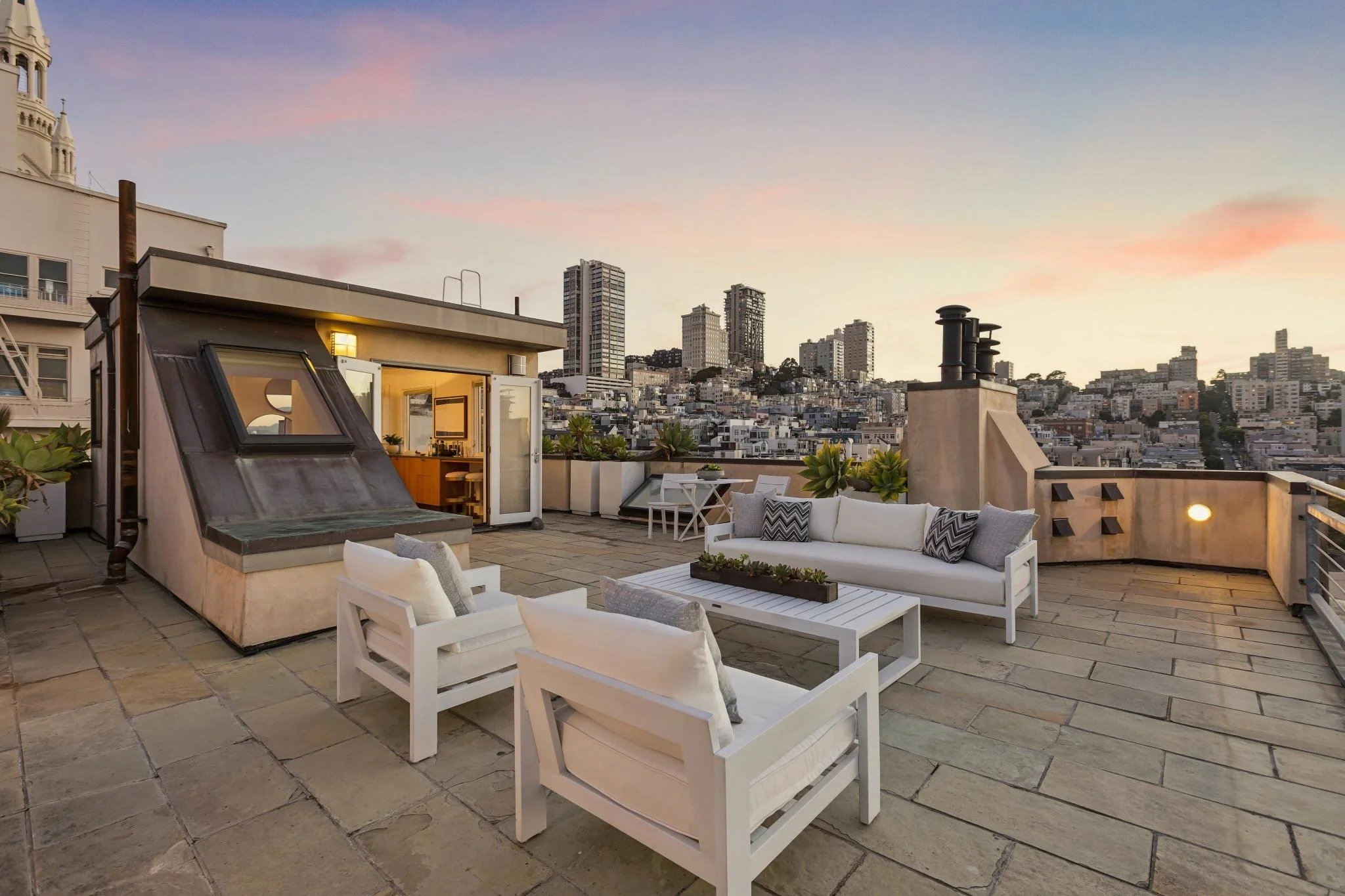 A rooftop patio at sunset with white outdoor furniture, potted plants, rooftop windows, and a city skyline in the background.