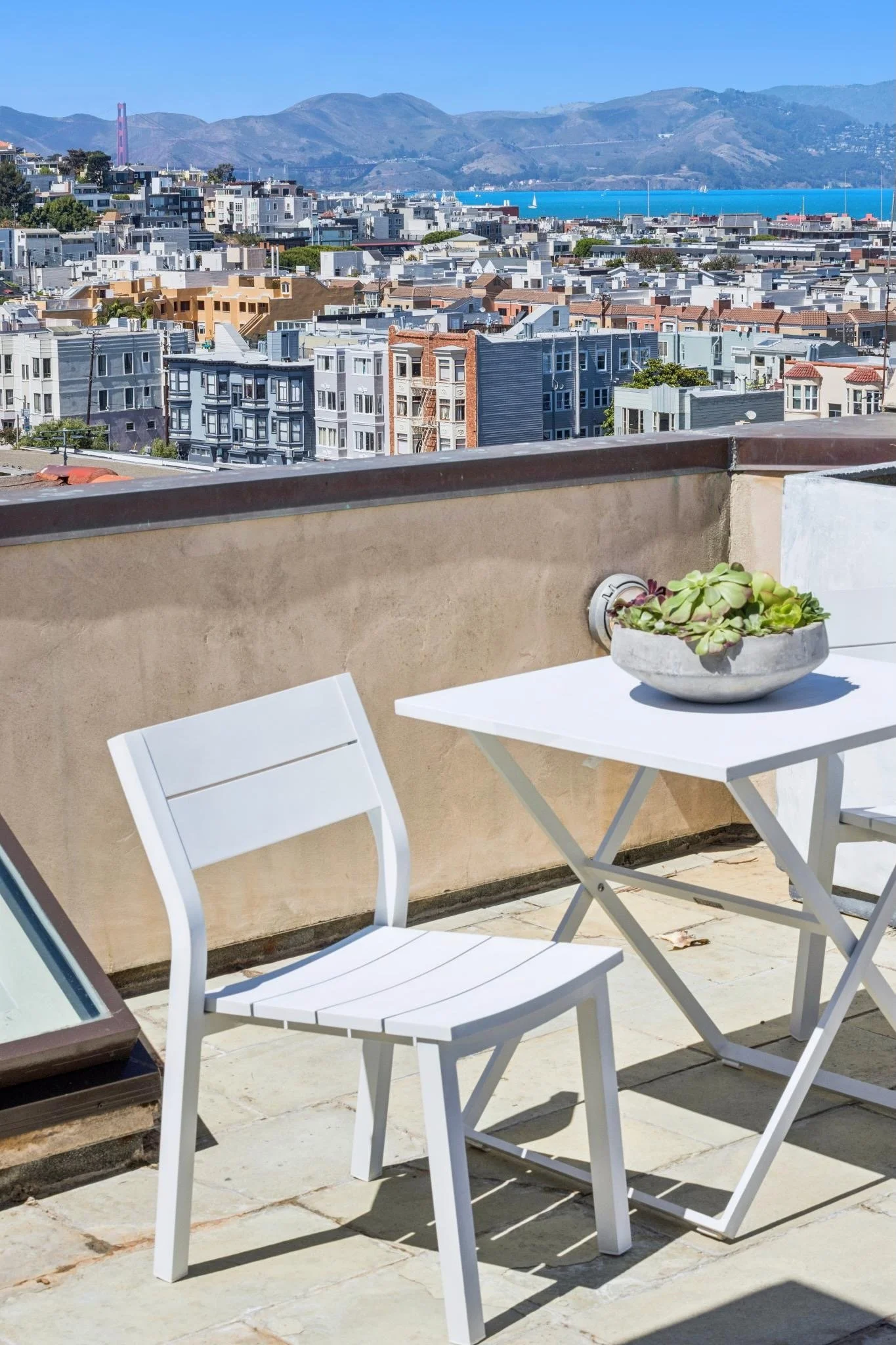 View from a terrace or balcony overlooking a cityscape with colorful buildings, hills, and water in the background, with a small table and chairs in the foreground.