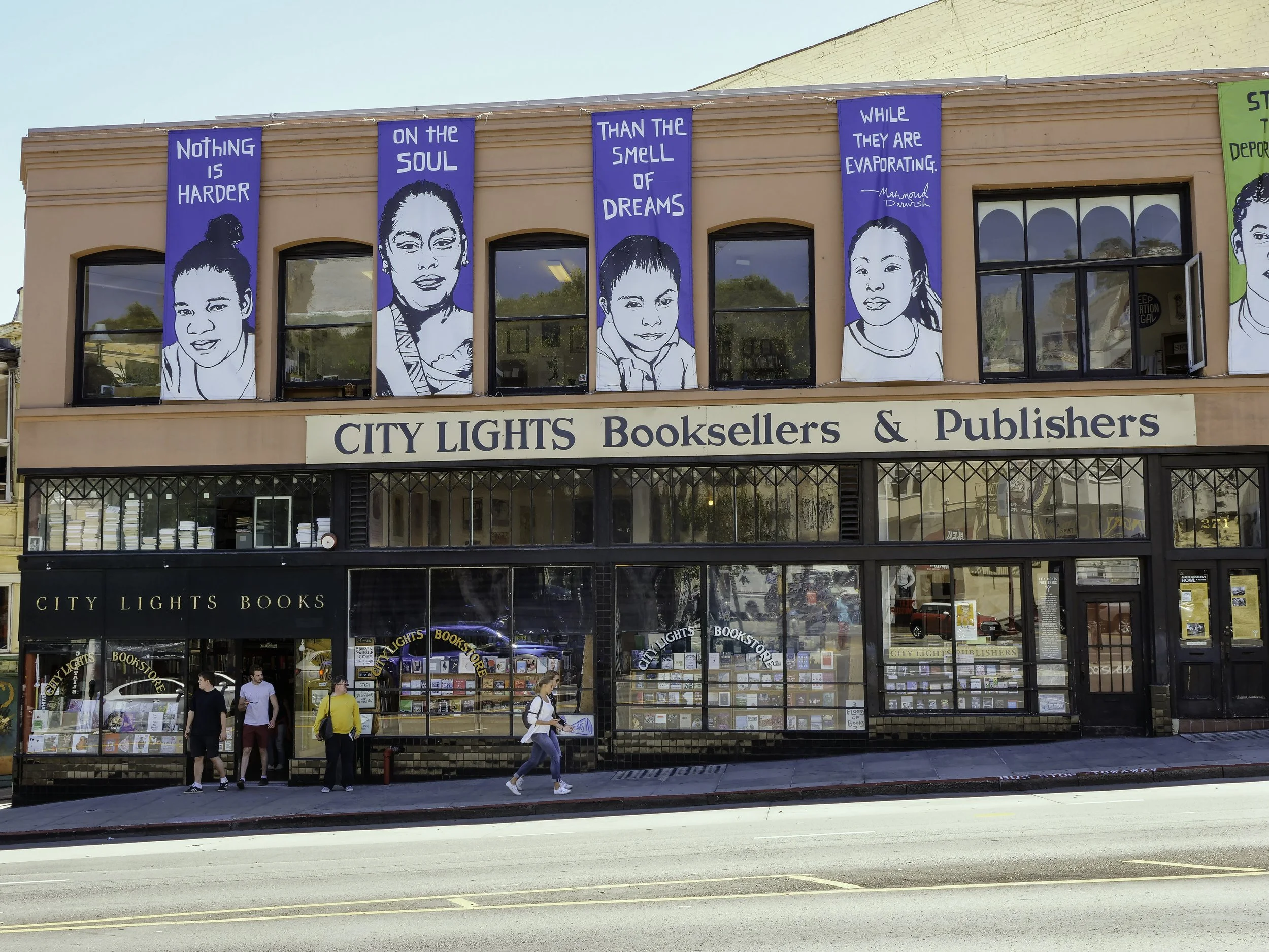 The exterior of a bookstore named City Lights Booksellers & Publishers, with large blue banners displaying quotes and artwork of women on the upper part of the building, and several people walking outside on the sidewalk.