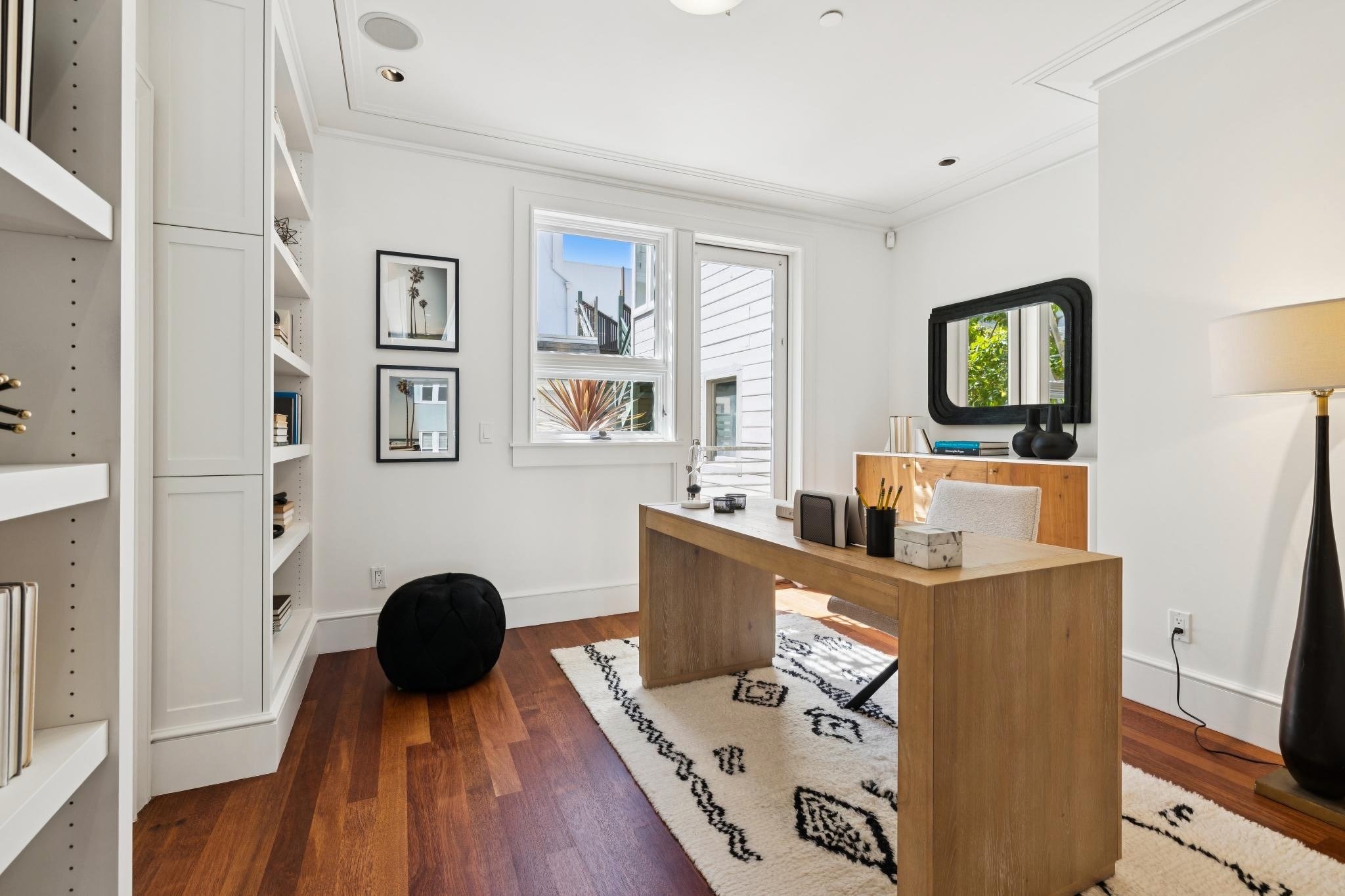 A home office with white walls, a wooden desk, a white bookshelf on the left, a window and a door On the back wall, black and white framed pictures of palm trees. A mirror on the right wall, black and white patterned rug, a black pouf, and a tall bla
