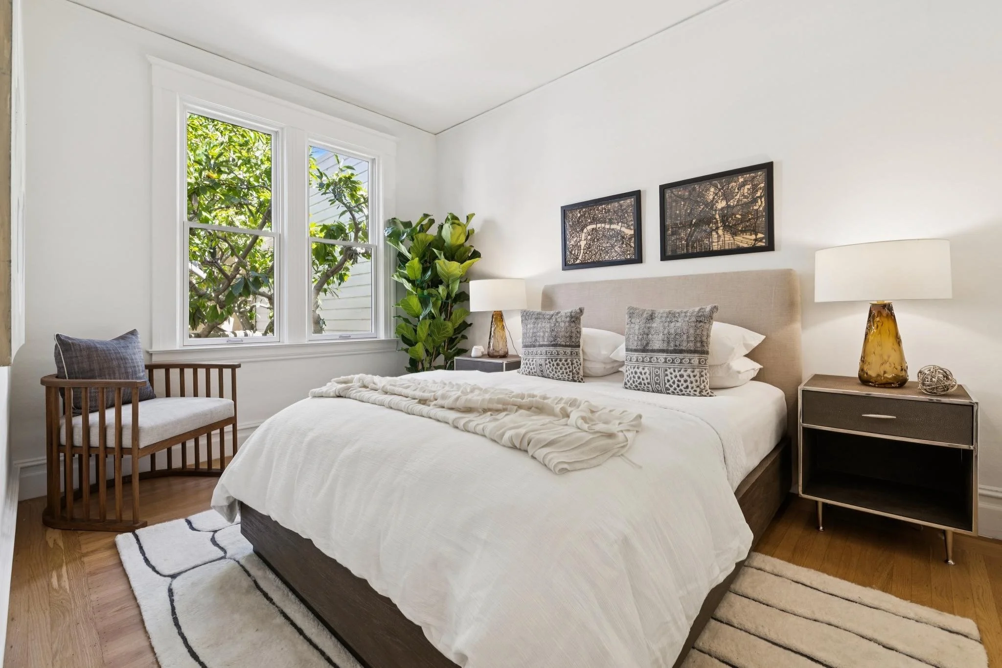 Bright bedroom with a large window showing green foliage outside. The bed has white bedding with decorative pillows, flanked by a nightstand with a amber-colored lamp and a modern painting on the wall. There's a wooden chair with a cushion near the w