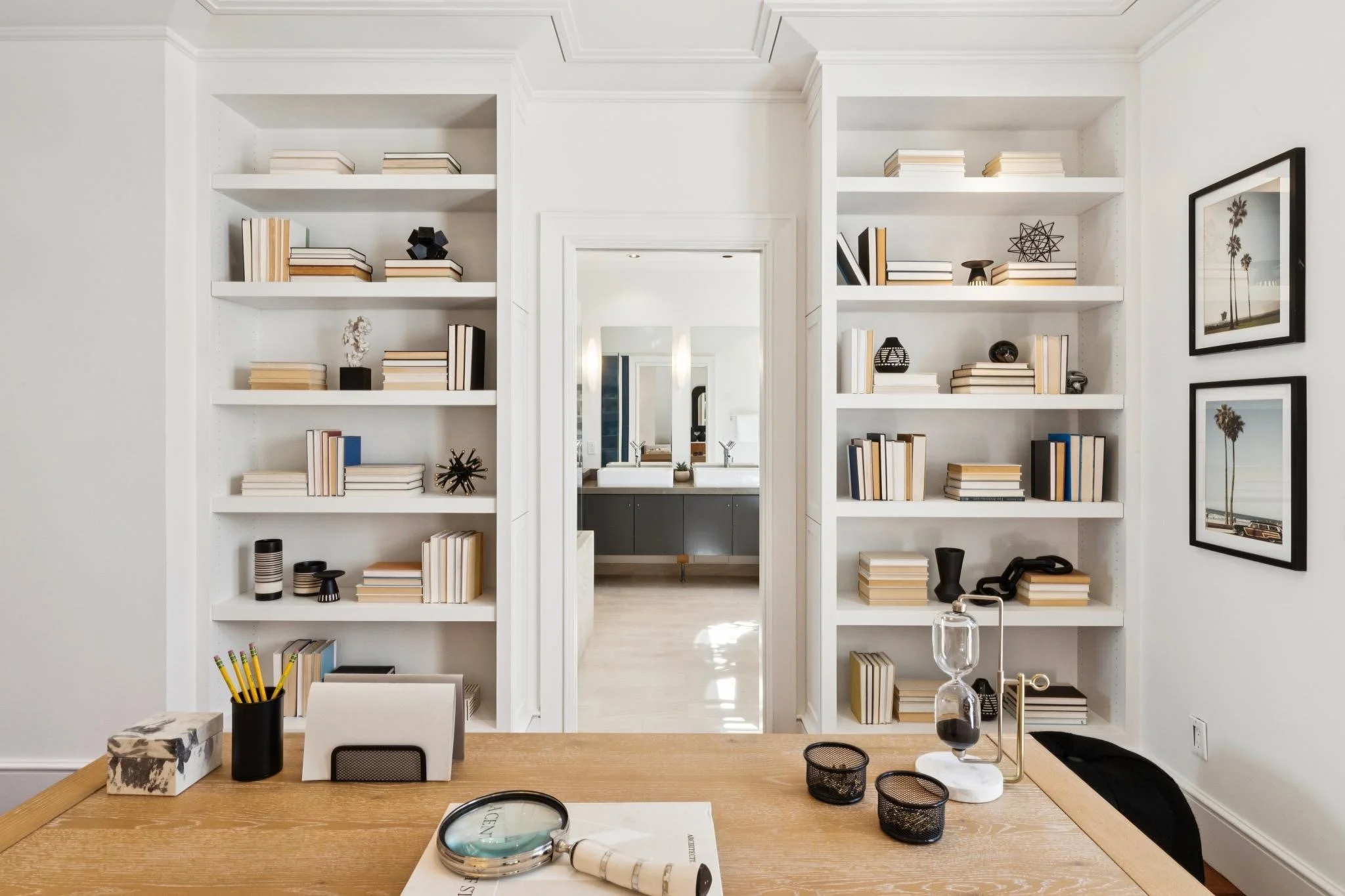 View of a modern interior room with white built-in bookshelves filled with books and decorative objects, leading to a bathroom with double sinks visible through an open doorway. A wooden table in the foreground holds various items including a clock, 