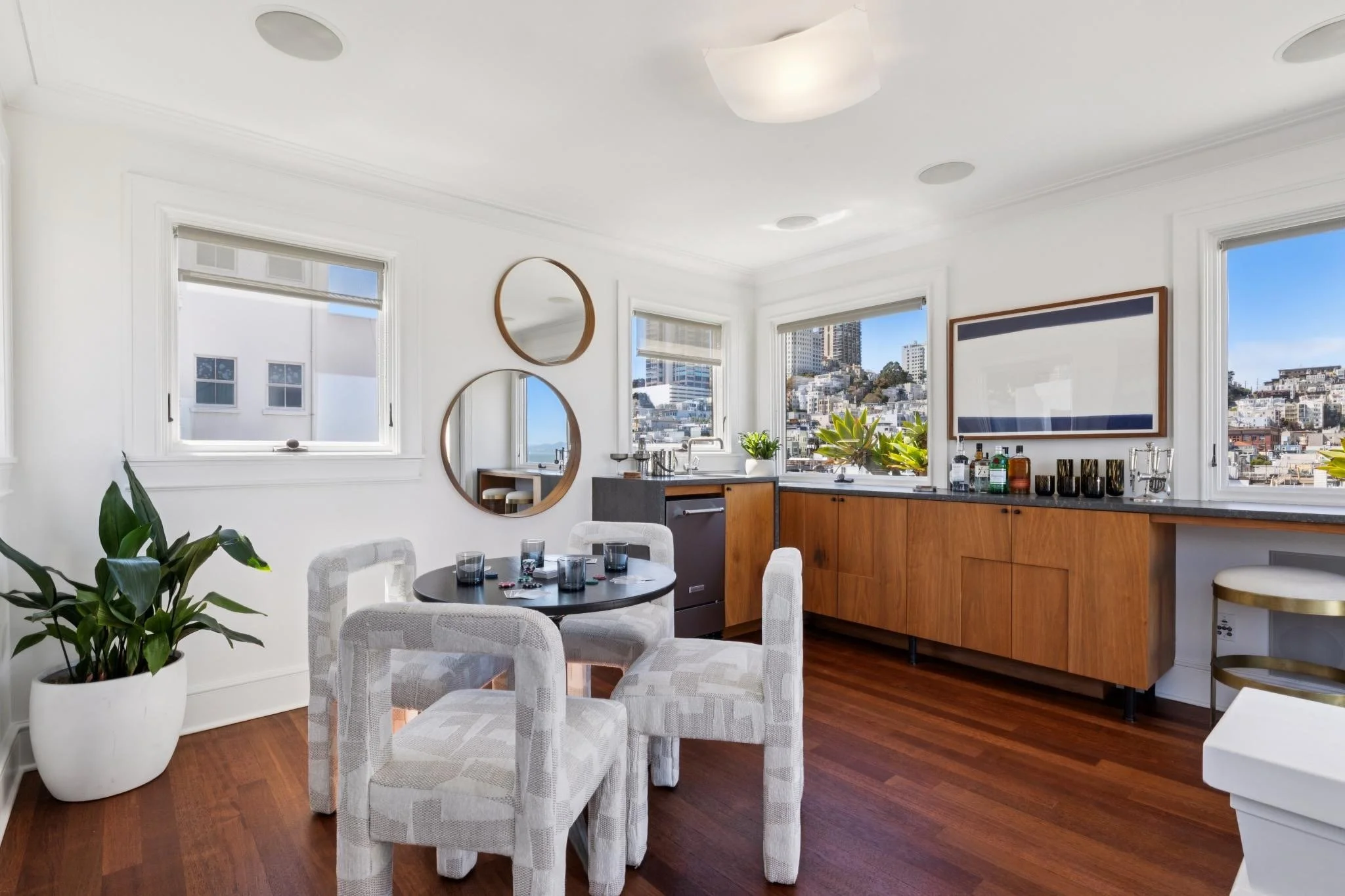 A modern kitchen and dining area with white walls, three windows showing a cityscape, wooden cabinetry, a black countertop, and round ceiling lights. A round mirror hangs on the wall, and there are white chairs around a black dining table set with gl
