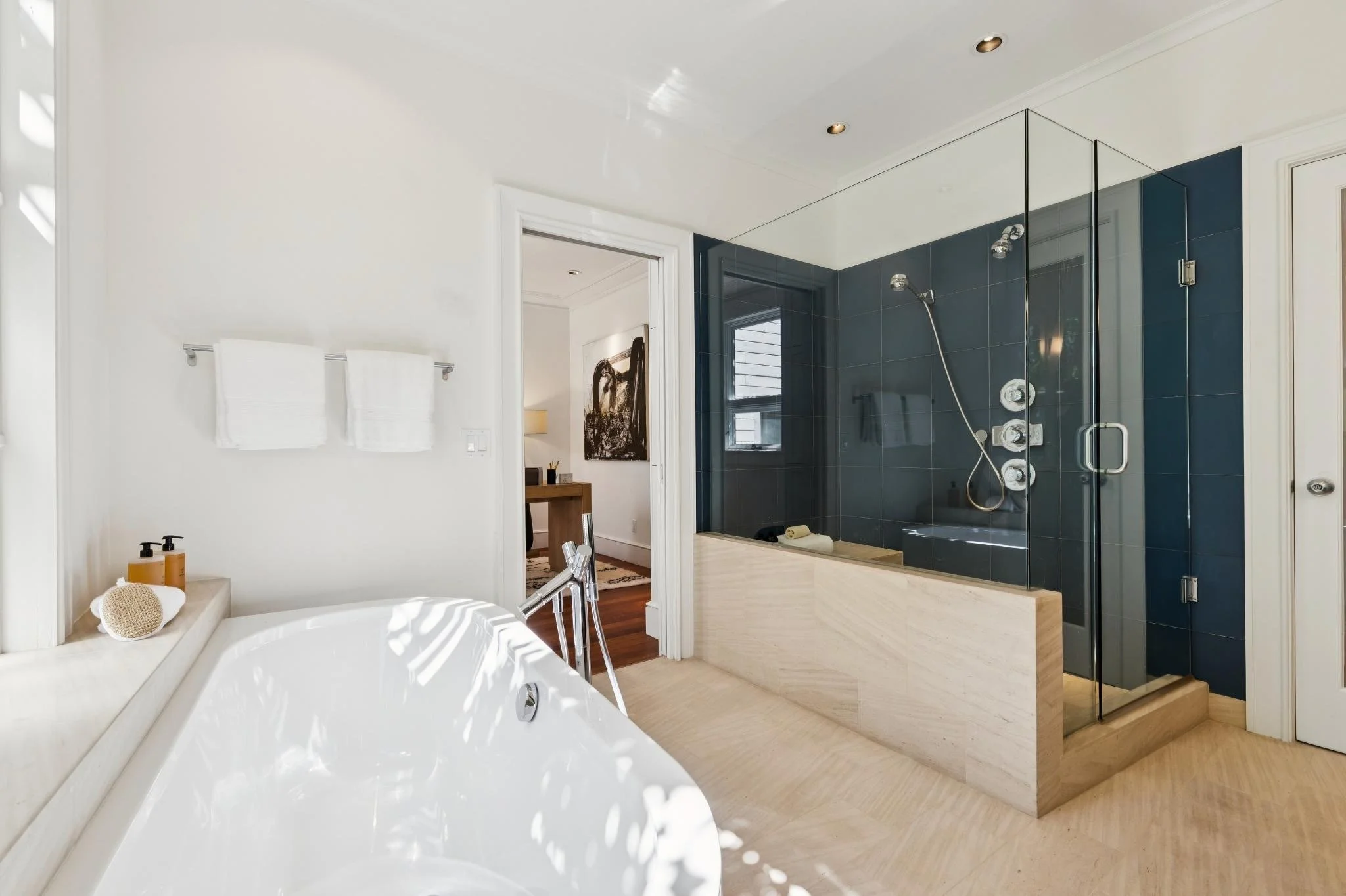 Modern bathroom with a white bathtub, a glass shower enclosure with dark tiles, and a doorway leading to a bedroom with a black and white painting on the wall.