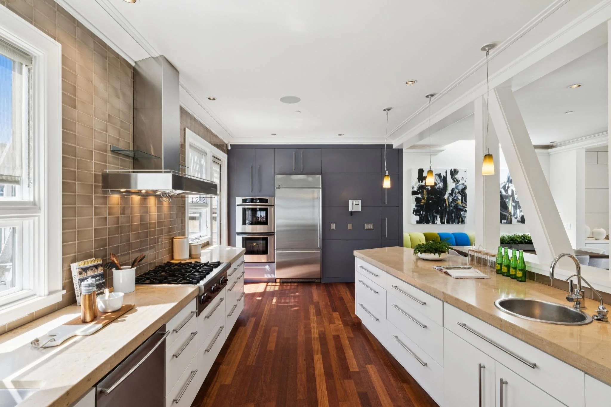Modern kitchen with white and gray cabinetry, a beige marble island, and stainless steel appliances, including a double oven and a large refrigerator. There are pendant lights hanging over the island and hardwood floors.