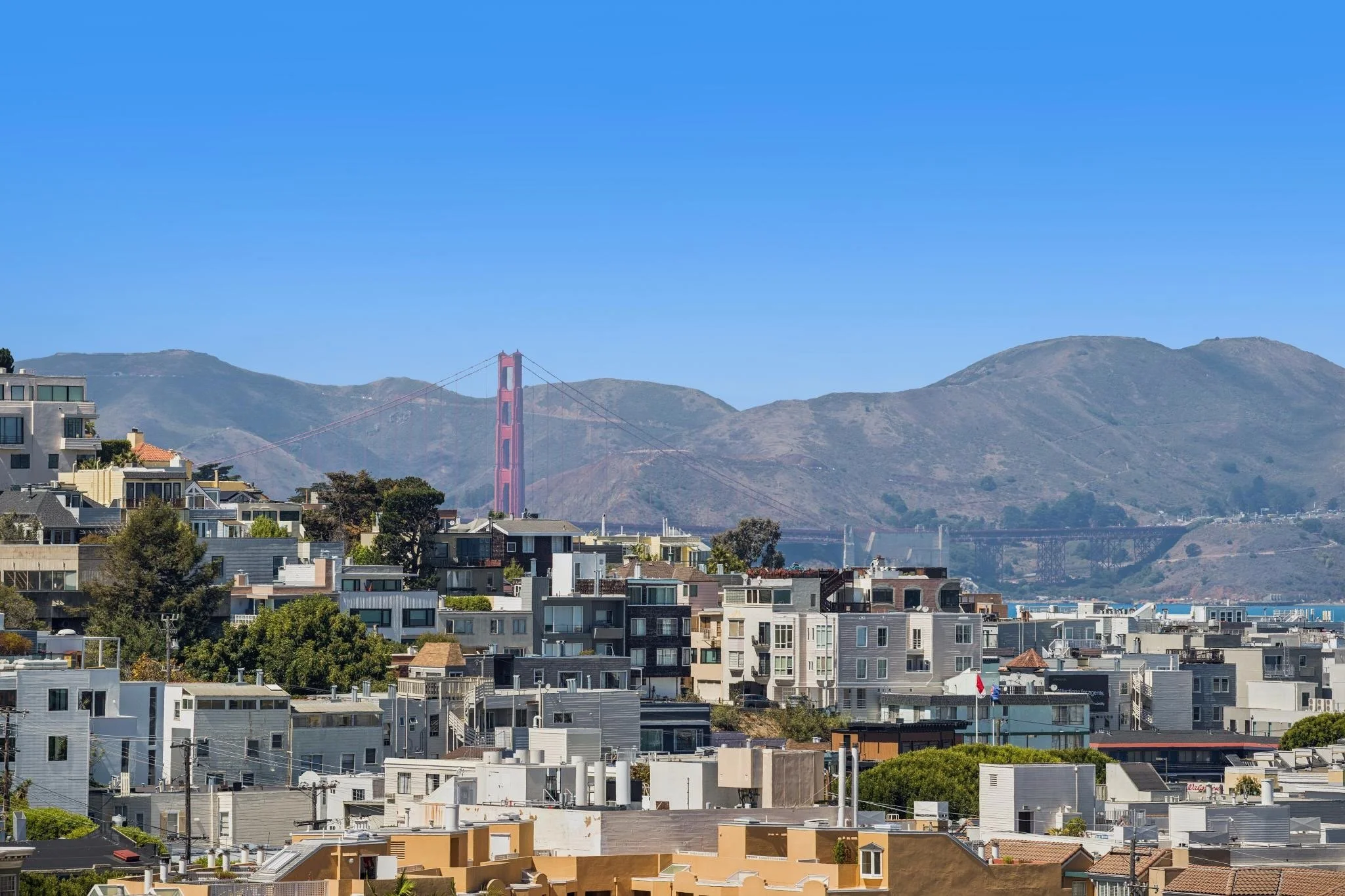 Cityscape of San Francisco with residential buildings in the foreground, the Golden Gate Bridge in the middle distance, and hills in the background under a clear blue sky.