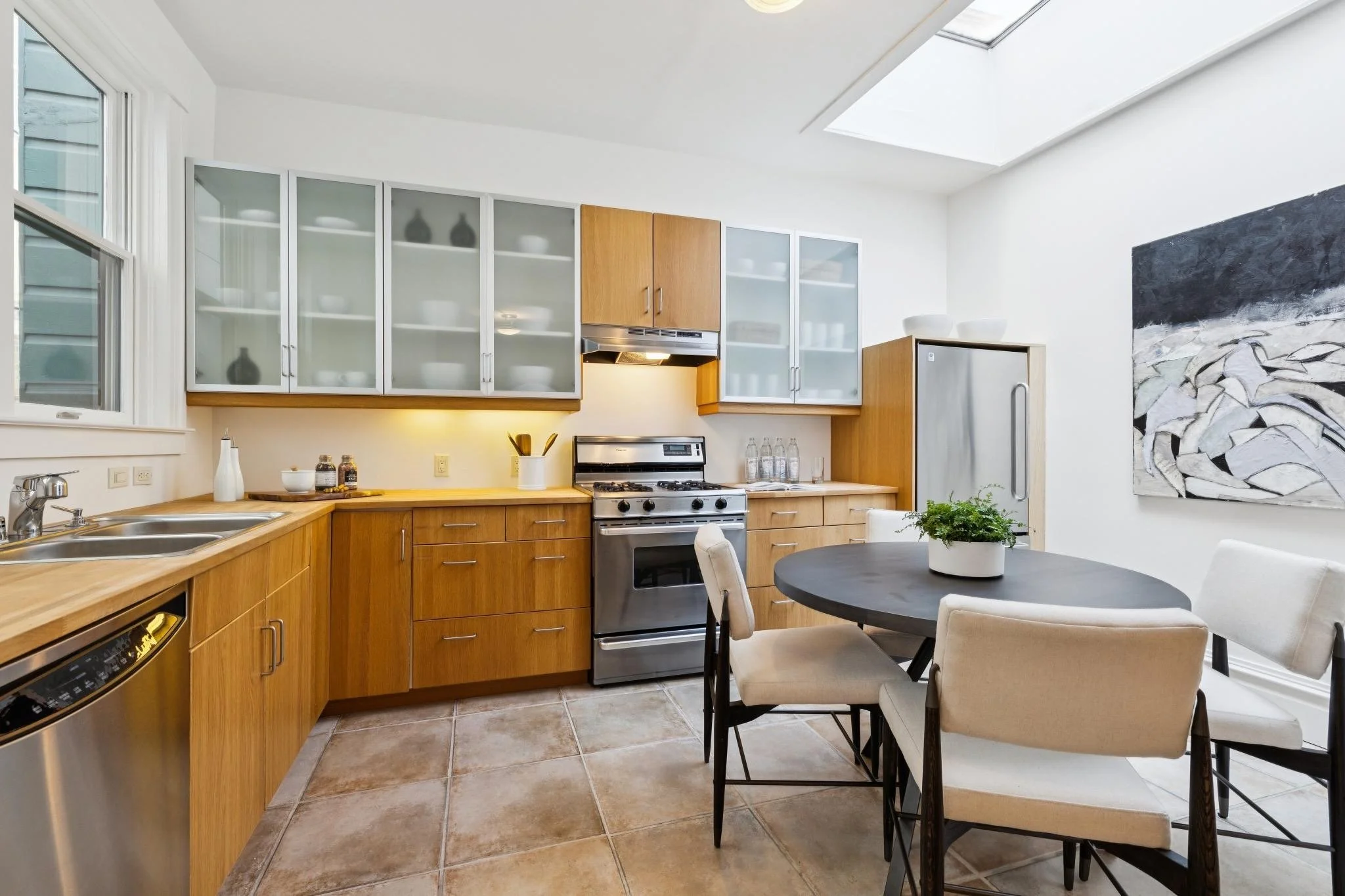 A modern kitchen with wooden cabinets, a stainless steel stove, and a table with four cream-colored chairs. There are glass-front cabinets, a window, and a large abstract artwork on the wall.