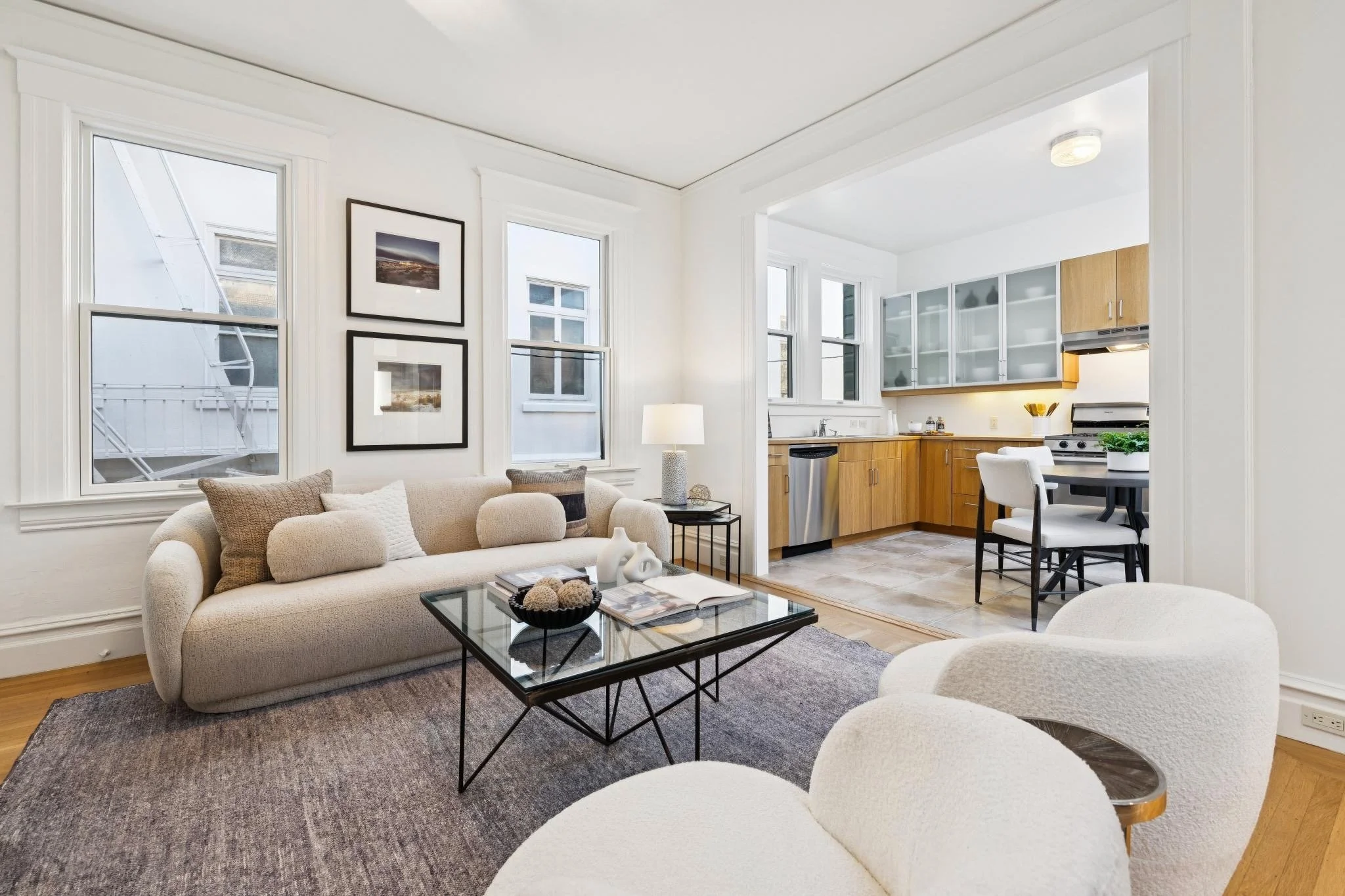 Living room with beige sofa, white chairs, black coffee table, artwork on white walls, and open kitchen with wooden cabinets and gray glass-front doors.