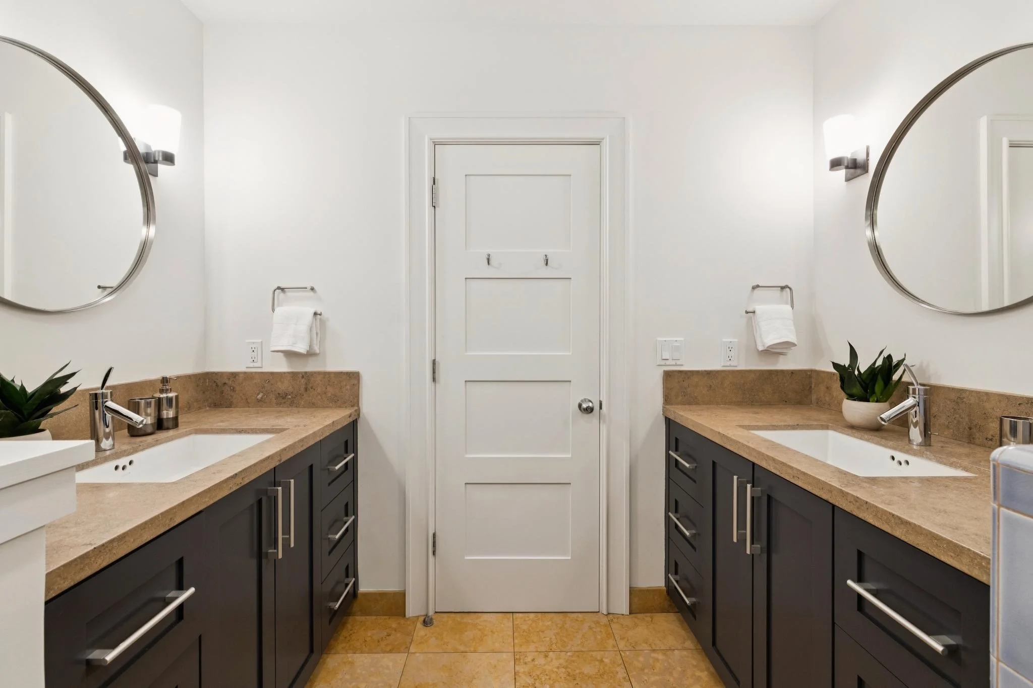 Double vanity bathroom with black cabinets, beige countertops, two oval mirrors, wall-mounted lights, and a white door in the center.