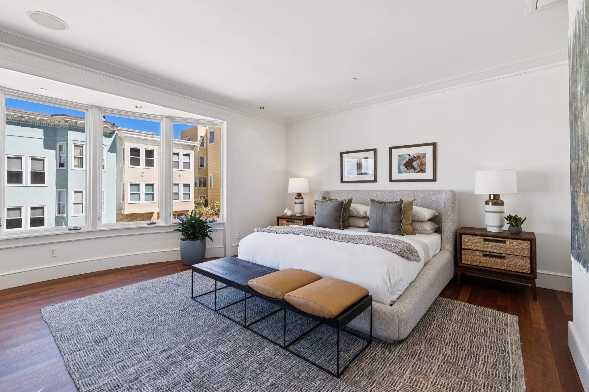 Bright bedroom with large window, gray upholstered bed with pillows, two matching nightstands with lamps, framed artwork on white walls, wooden floor, and a patterned rug.