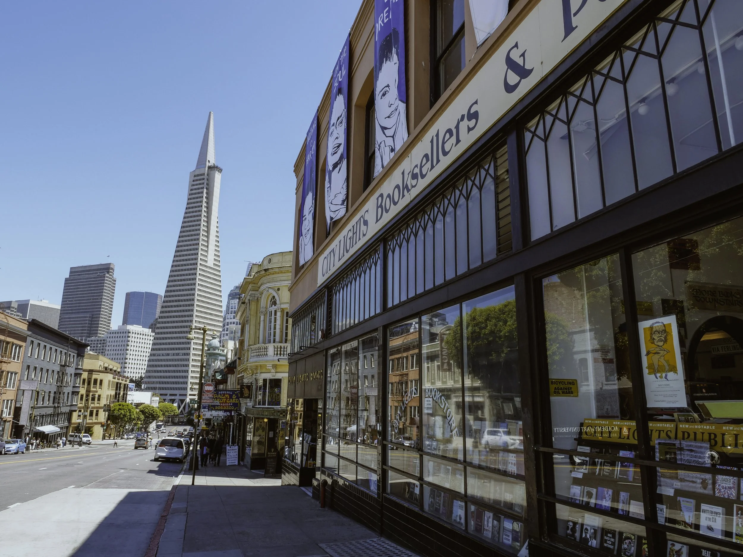 City street view with a bookstore on the right side of the image, featuring large posters of comics on the upper windows. Modern skyscrapers, including the Transamerica Pyramid, are visible in the background under a clear blue sky.
