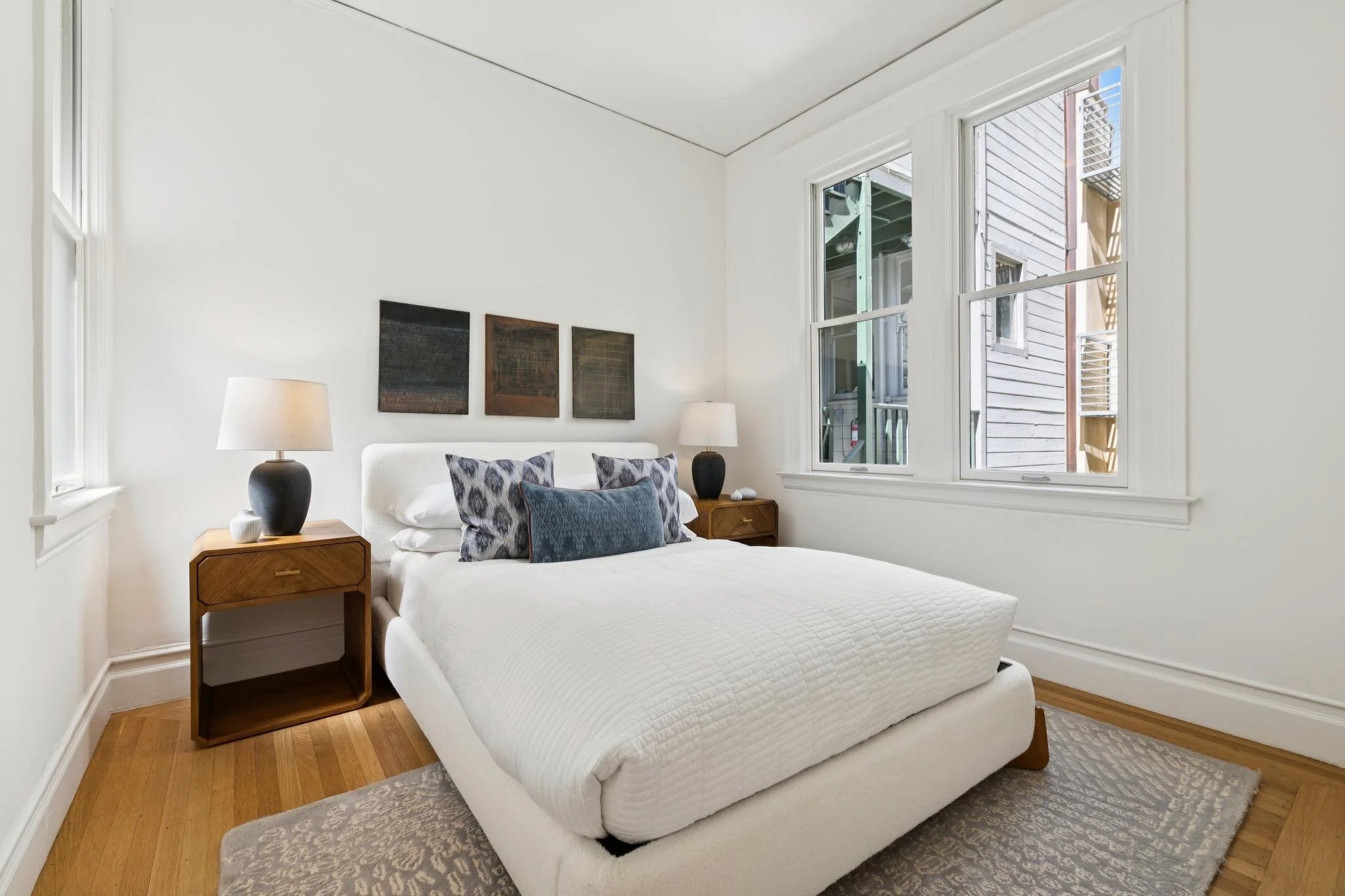 A bedroom with a white bed, three decorative pillows, two bedside tables with black lamps, three paintings above the bed, a window with view of the outside, and a rug on a wooden floor.
