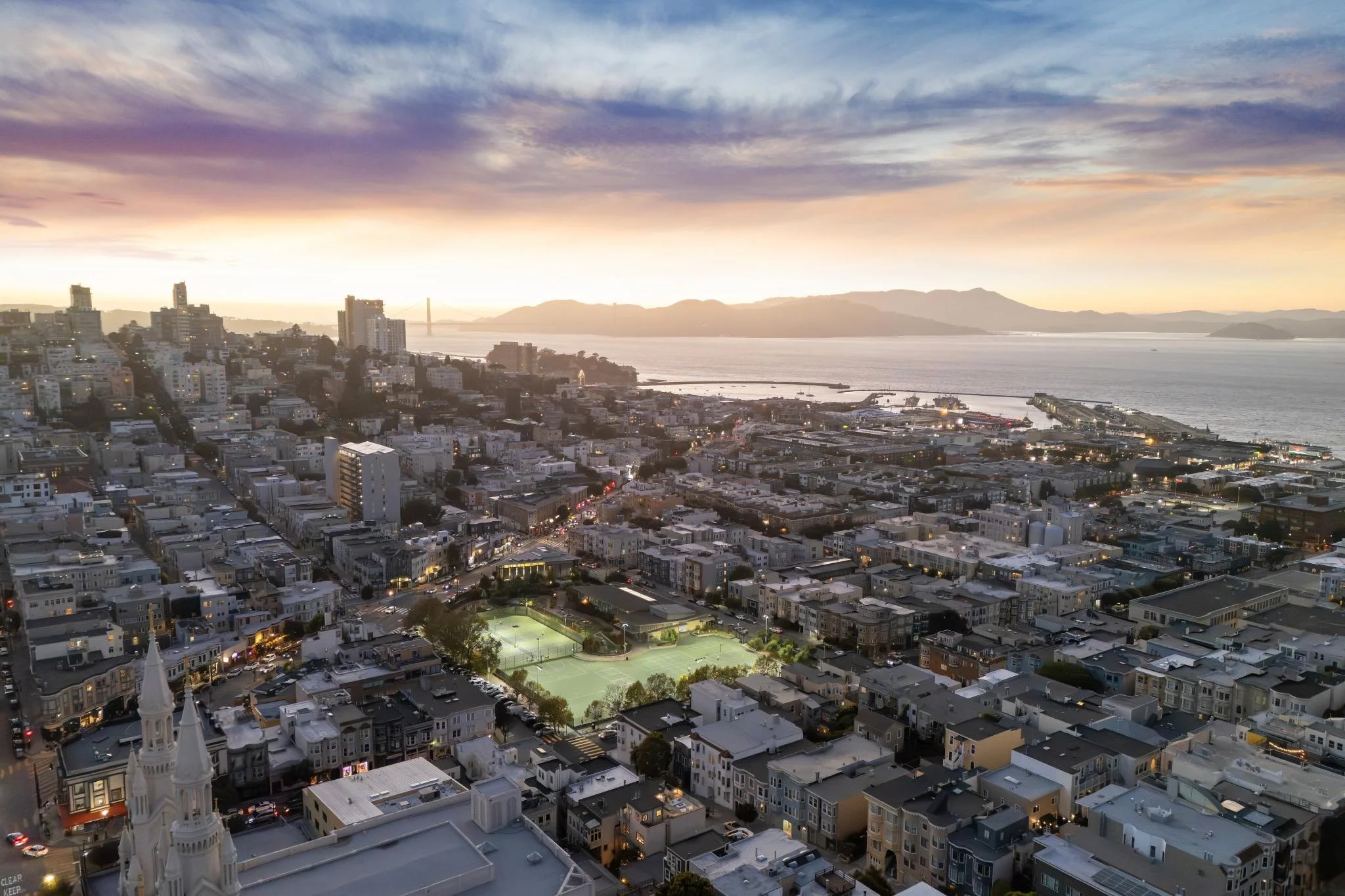 A cityscape view of San Francisco during sunset with buildings, a tennis court, water, and hills in the background.