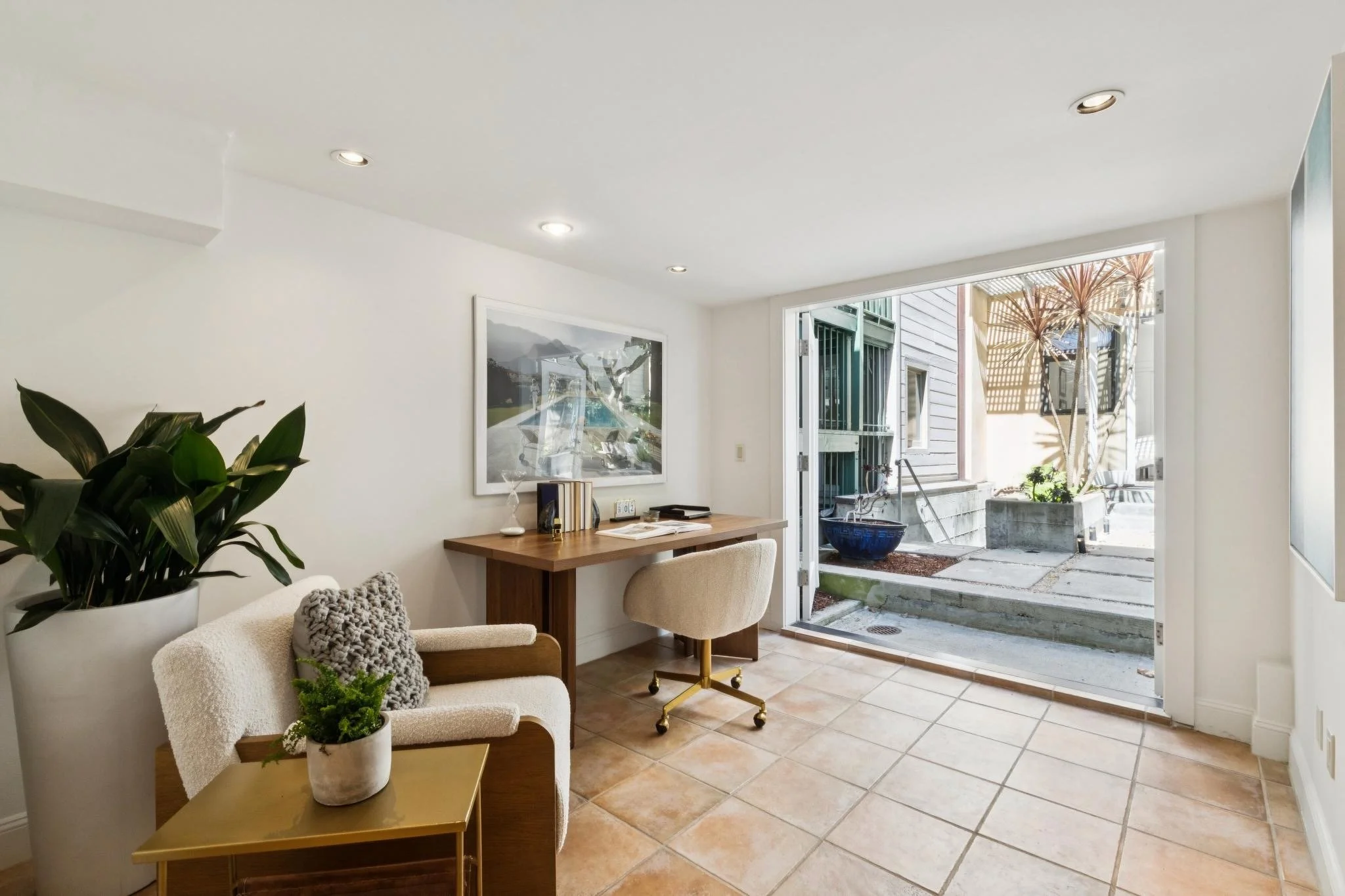 A cozy living room with a white couch decorated with knitted pillows, a small side table with potted plants, a wooden desk with a plant and books, a beige chair, and an open door leading to a patio with outdoor plants and sunlight.