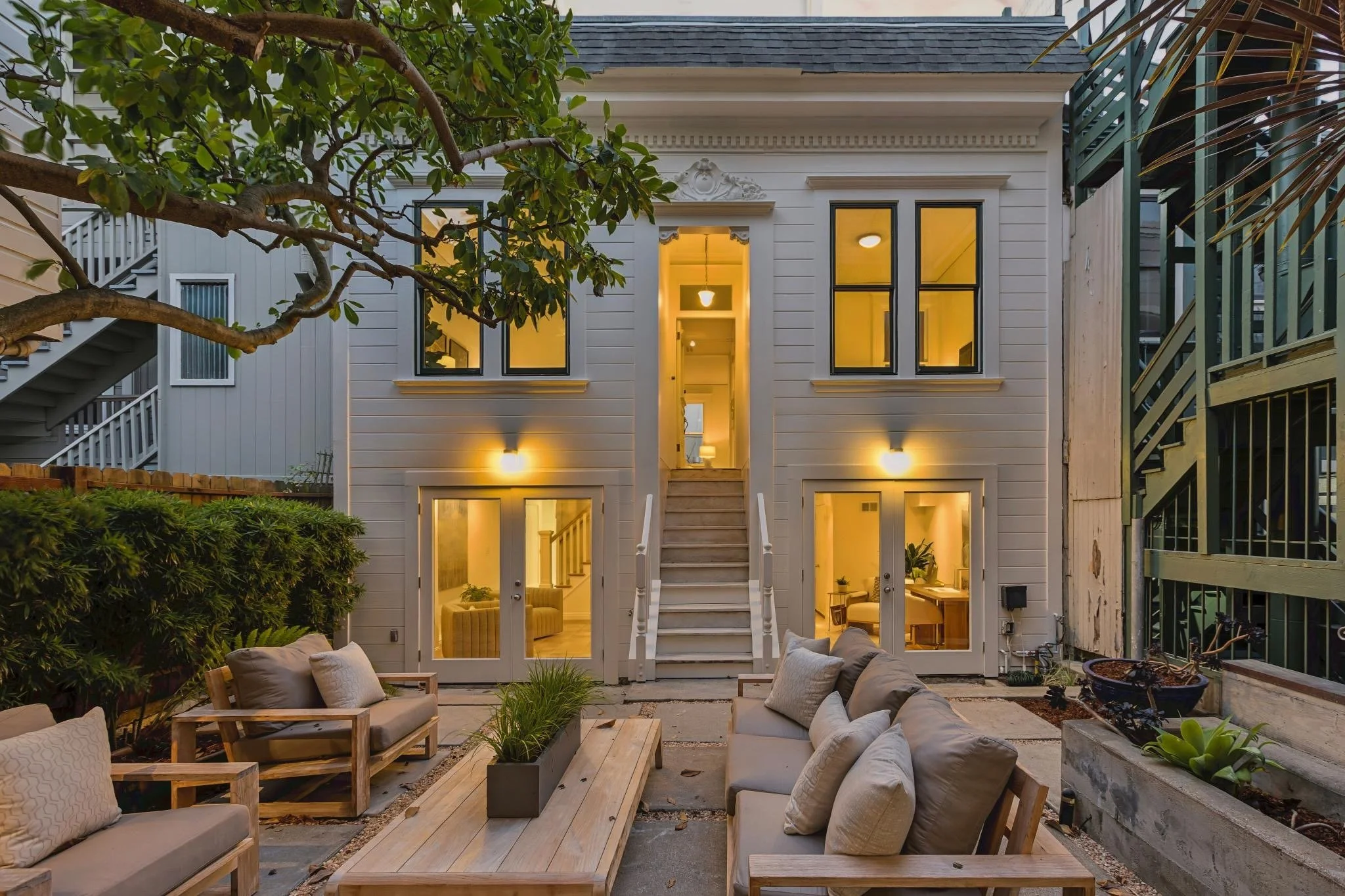 Backyard patio with beige outdoor furniture, a wooden coffee table, surrounded by green plants, trees, and shrubbery, with a white multi-story house with lit interior windows in the background.