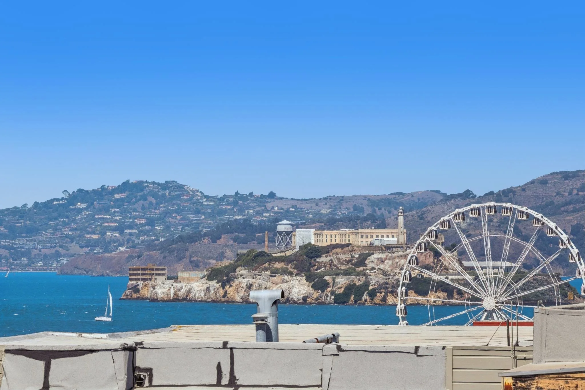 View of the San Francisco Bay with a Ferris wheel, sailboats, and a hillside with buildings and trees under a clear blue sky.