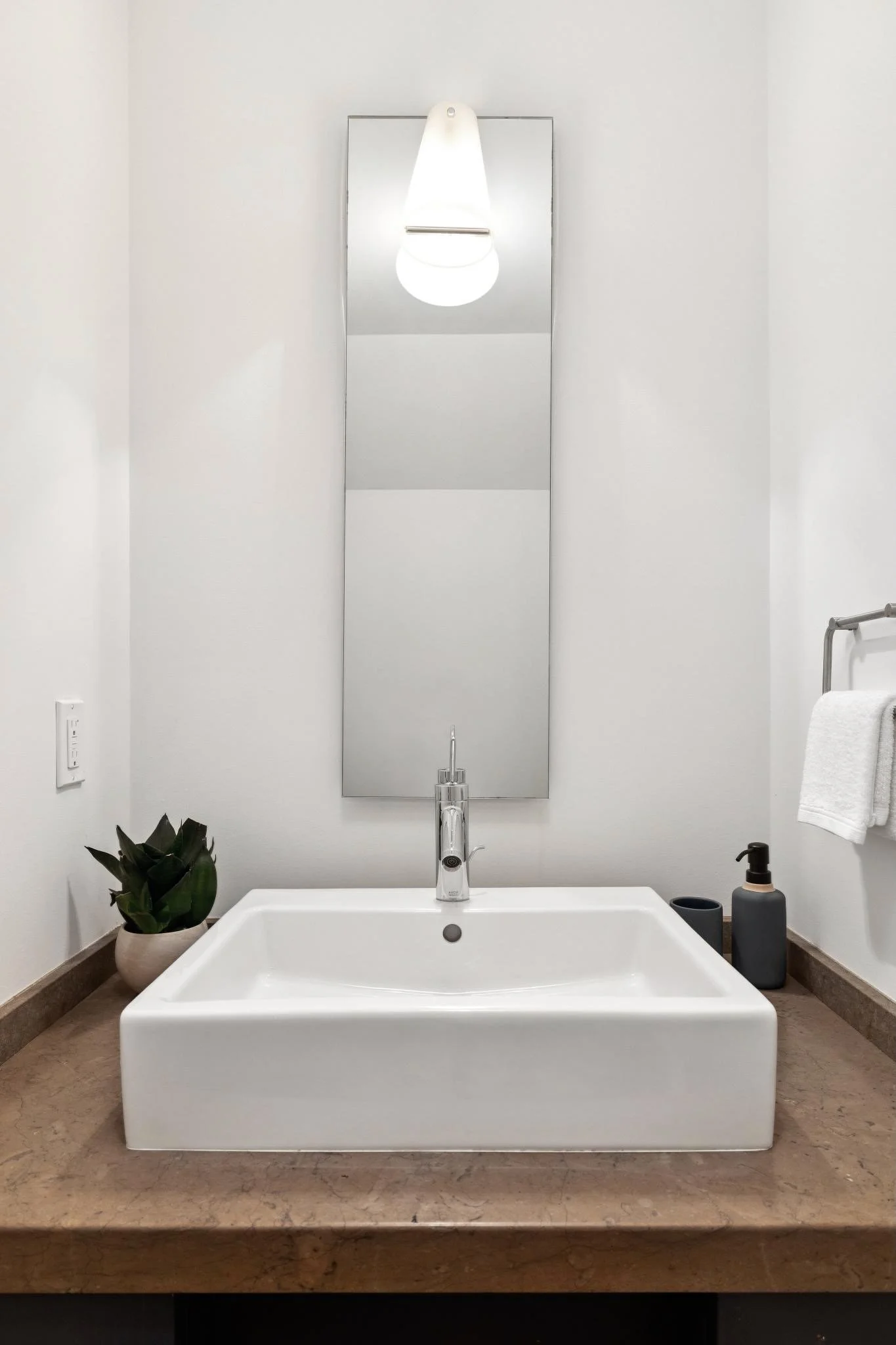 Modern bathroom with a white sink, a faucet, a mirror, a lamp, a potted plant, a soap dispenser, and a towel on a rack.