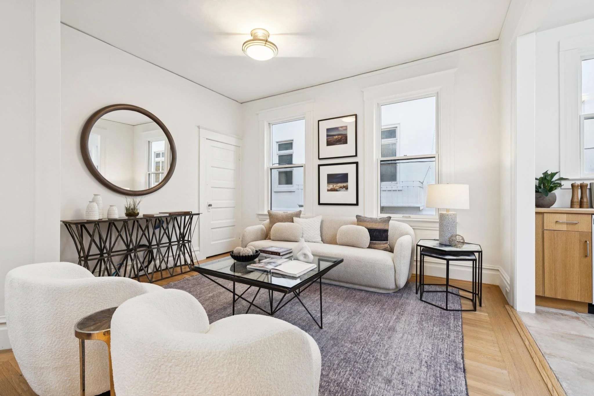 Bright living room with white walls, multiple windows, a beige sofa, white armchair, black coffee table, side table with a lamp, wall art, decorative vases, mirror, and plants.