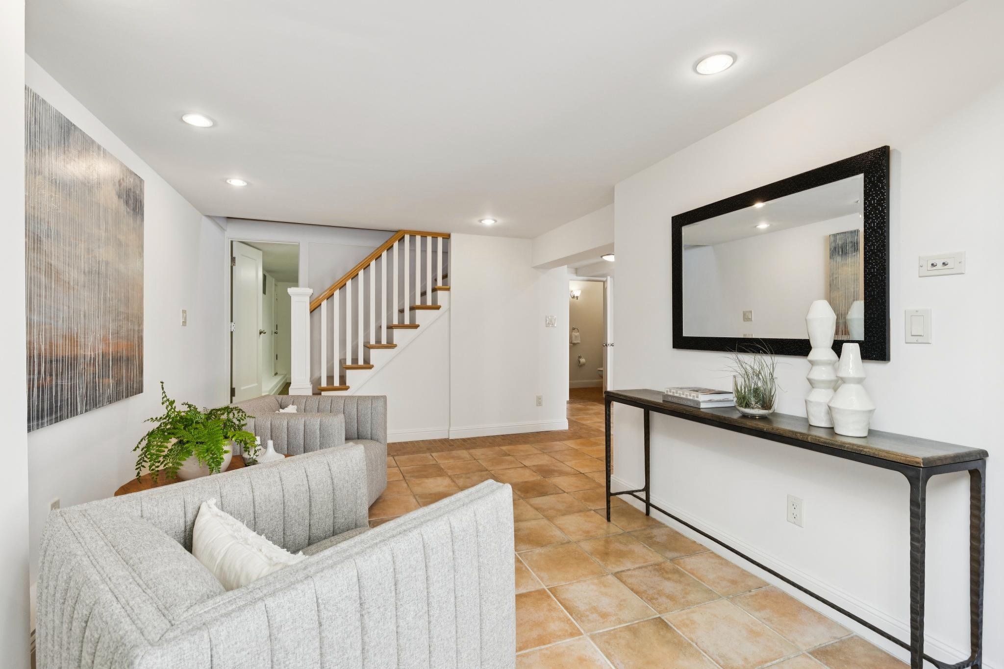 Living room with white walls, terracotta tile flooring, gray sofas, a large black-framed mirror, a console table with white vases, and a staircase with a wooden handrail.