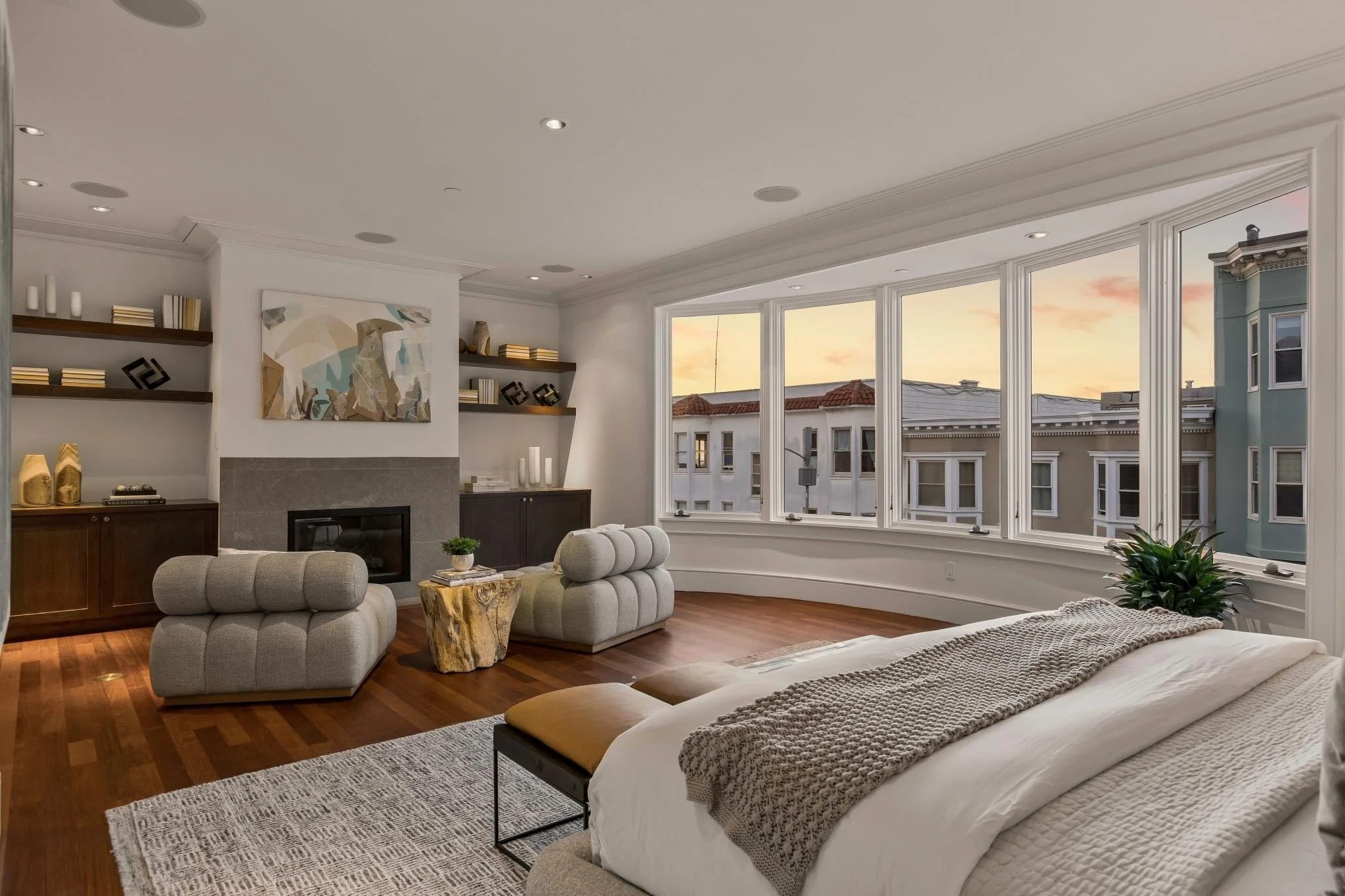 Living room with large bay window, beige sofas, abstract wall art, wooden shelves with books and decor, hardwood floors, and sunset view outside.
