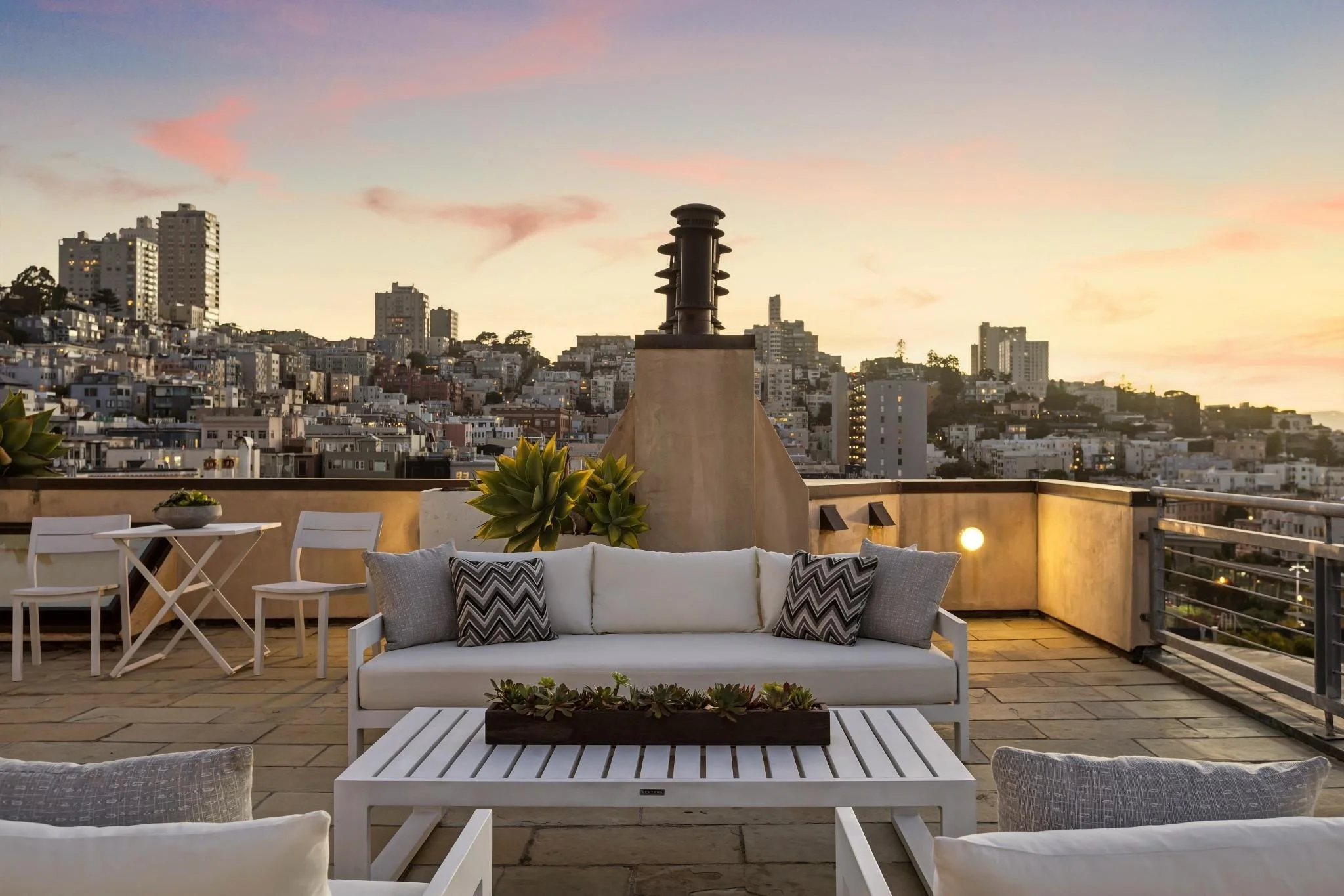 A rooftop terrace with white outdoor furniture, including a sofa with pillows, a coffee table, and chairs, overlooking a cityscape at sunset with high-rise buildings and a chimney stack in the background.