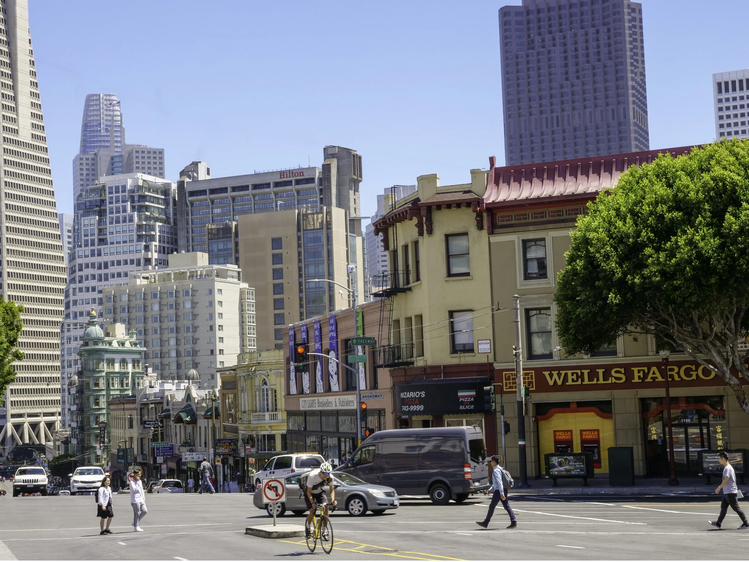A busy city street scene with pedestrians crossing the road, cars driving, and tall buildings in the background. There are signs for Wells Fargo, pizza, and other businesses, and a person riding a bike.