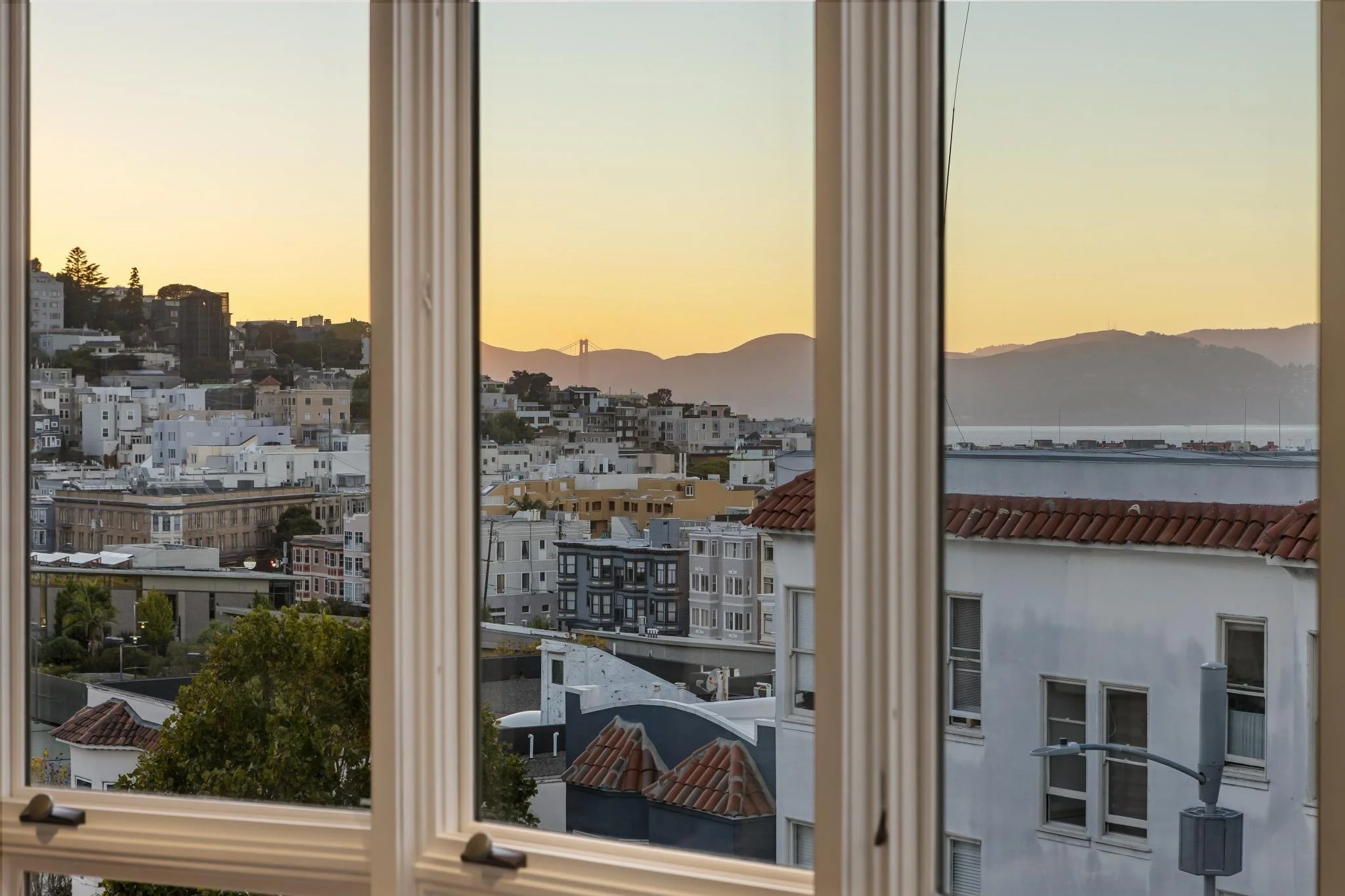 View of a cityscape through a window with beige blinds, showing residential buildings, trees, hills, and a body of water during sunset.