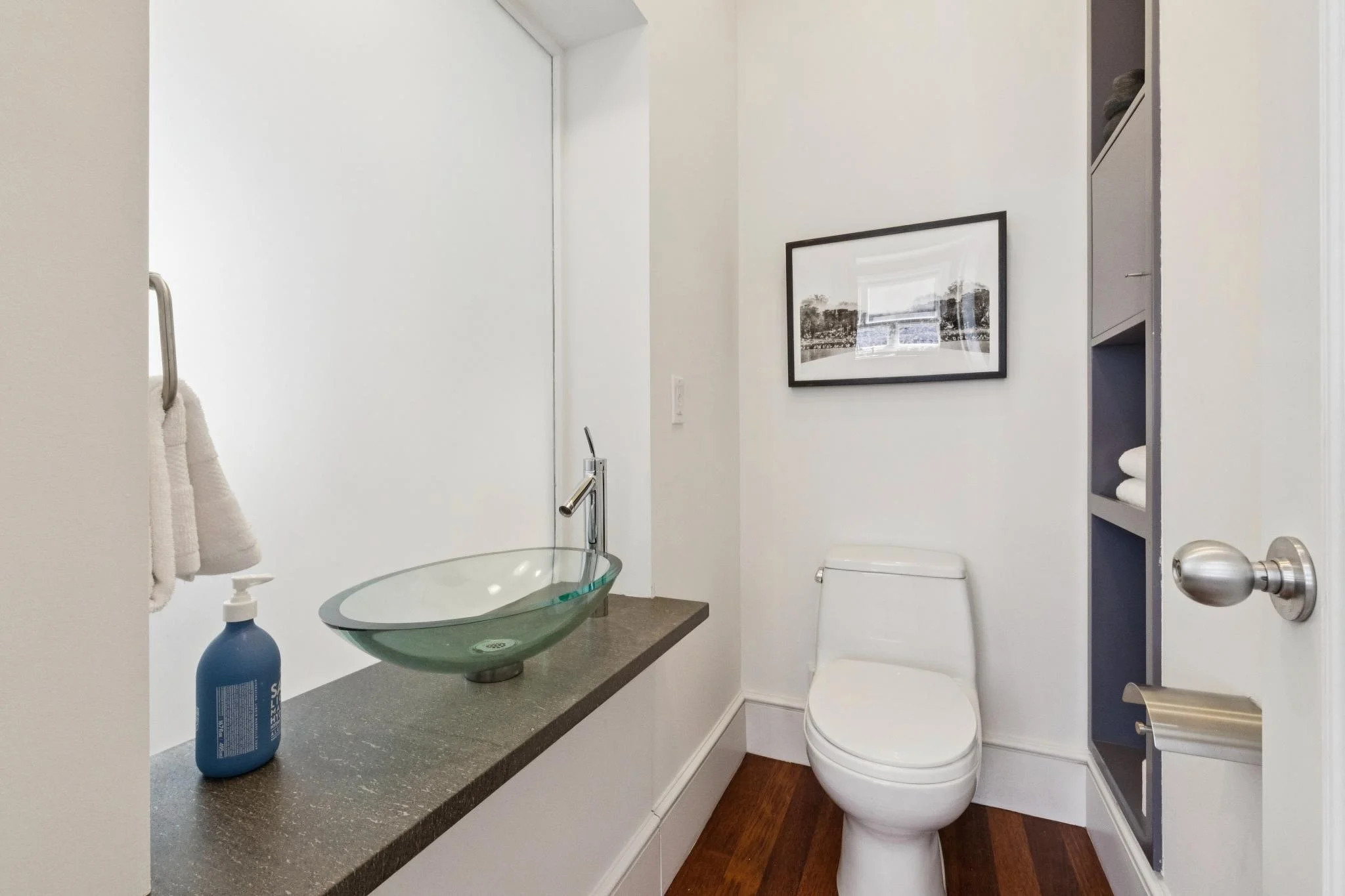 A modern bathroom with a white toilet, a glass vessel sink on a gray countertop, a framed black-and-white landscape picture on the wall, and a tall built-in shelf with folded towels. There is a bottle of soap or sanitizer on the countertop and a towe