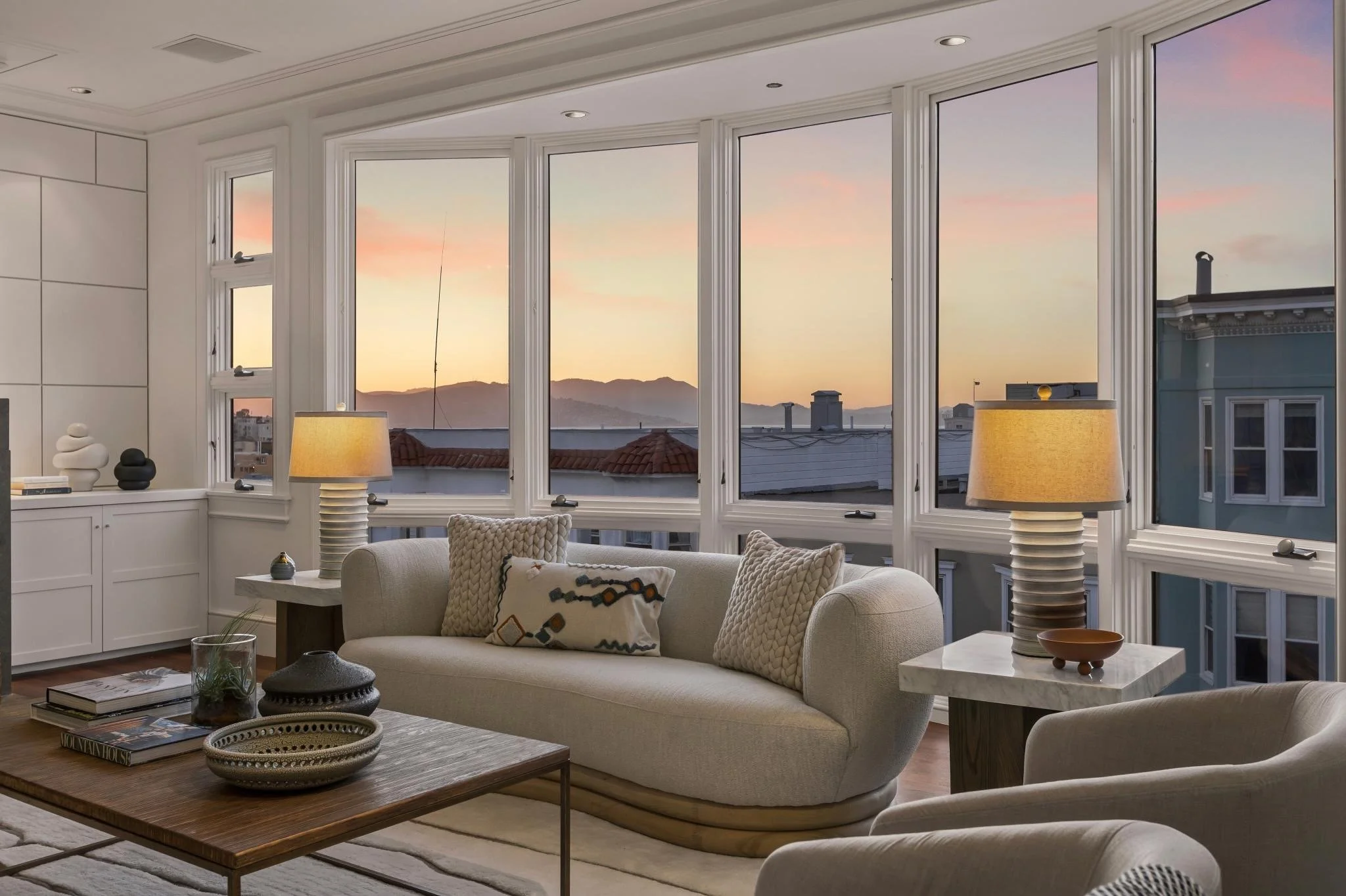 Living room with large windows showing a sunset view of mountains, with a sofa, coffee table, and lamps.
